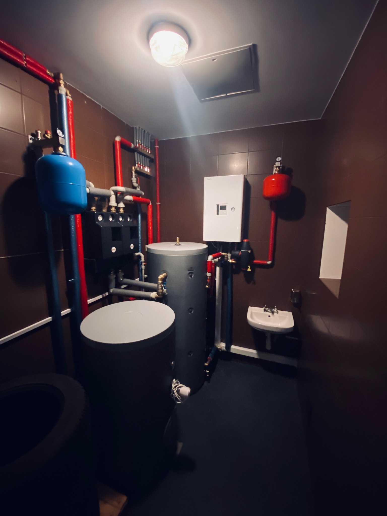 Zestaw instalacji grzewczej w kotłowni: dwa zbiorniki, biały piec, czerwone i niebieskie rury, mała umywalka. Brązowe płytki na ścianach, jasne światło sufitowe.