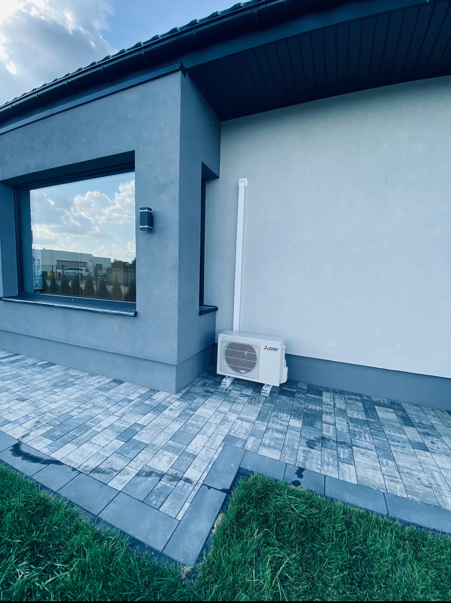 Nowoczesny budynek z zamontowaną jednostką zewnętrzną klimatyzacji Mitsubishi Electric, widoczną na tle szarej elewacji i kostki brukowej.