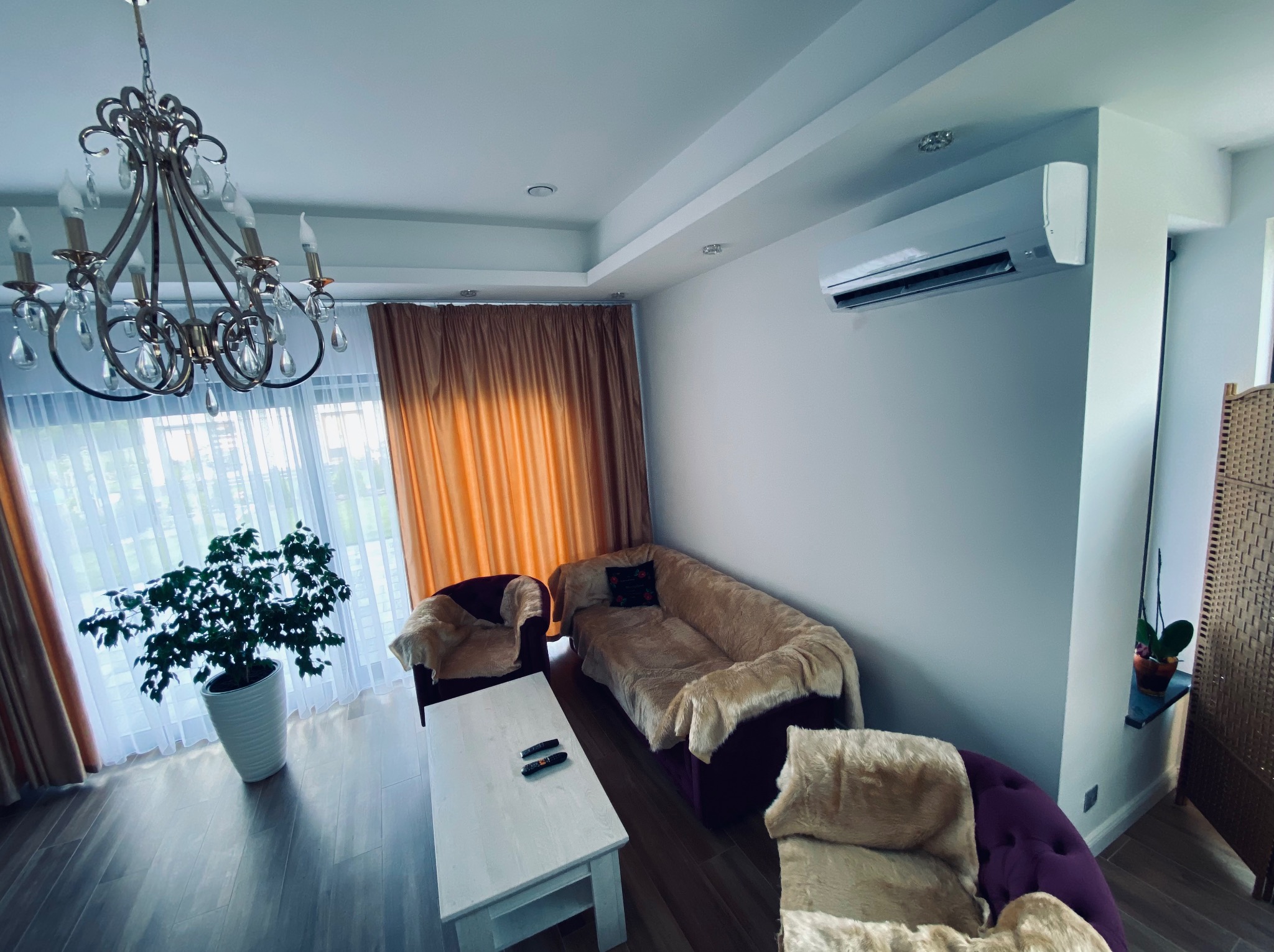 Nowoczesny salon z klimatyzacją ścienną, eleganckim żyrandolem i rośliną doniczkową. Widok na sofę, fotel i stolik kawowy. Jasne wnętrze z zasłonami w tle.