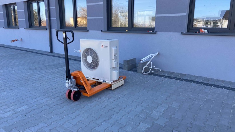 Biała jednostka zewnętrzna klimatyzacji Mitsubishi na pomarańczowym wózku paletowym przed szarym budynkiem z oknami. Widoczne przewody i bloczki betonowe.