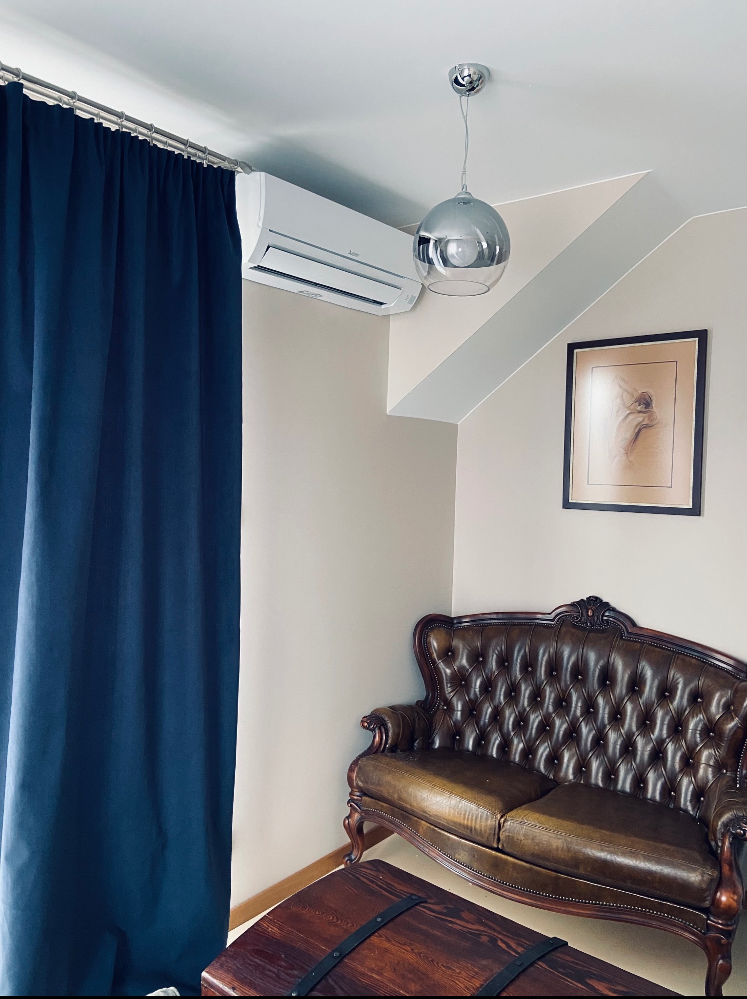 Klimatyzator ścienny Mitsubishi w eleganckim wnętrzu z ciemnoniebieską zasłoną, skórzaną sofą i lampą wiszącą. Ujęcie z dołu, podkreślające nowoczesność i komfort.