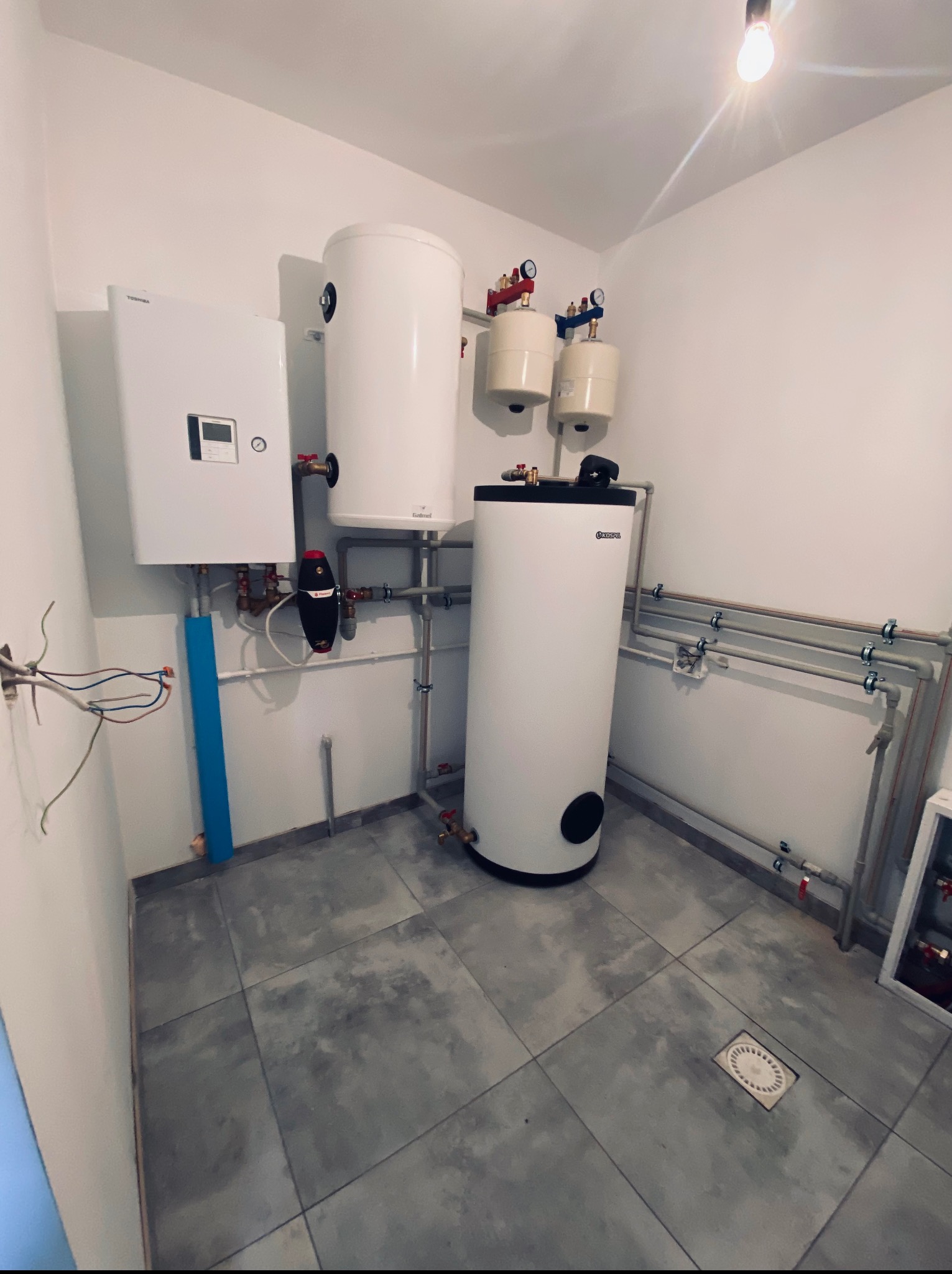 Nowoczesna kotłownia z białym piecem gazowym, zbiornikami i instalacją grzewczą. Szare płytki podłogowe, minimalistyczny design. Widoczne elementy instalacji.