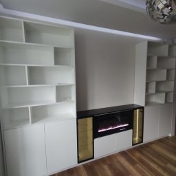 MALWA MEBLE Dariusz Jewulski - Biała zabudowa meblowa na ścianie z elektrycznym kominkiem i półkami. Nowoczesny design, minimalistyczny styl. Oświetlenie LED podkreśla detale.
