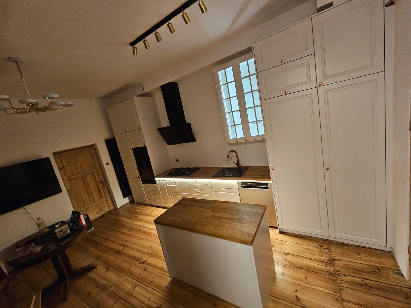 Jasna kuchnia z wyspą i drewnianą podłogą. Białe szafki z miedzianymi uchwytami, czarny okap i oświetlenie punktowe. Ustawiona pod kątem, widoczny fragment stołu.