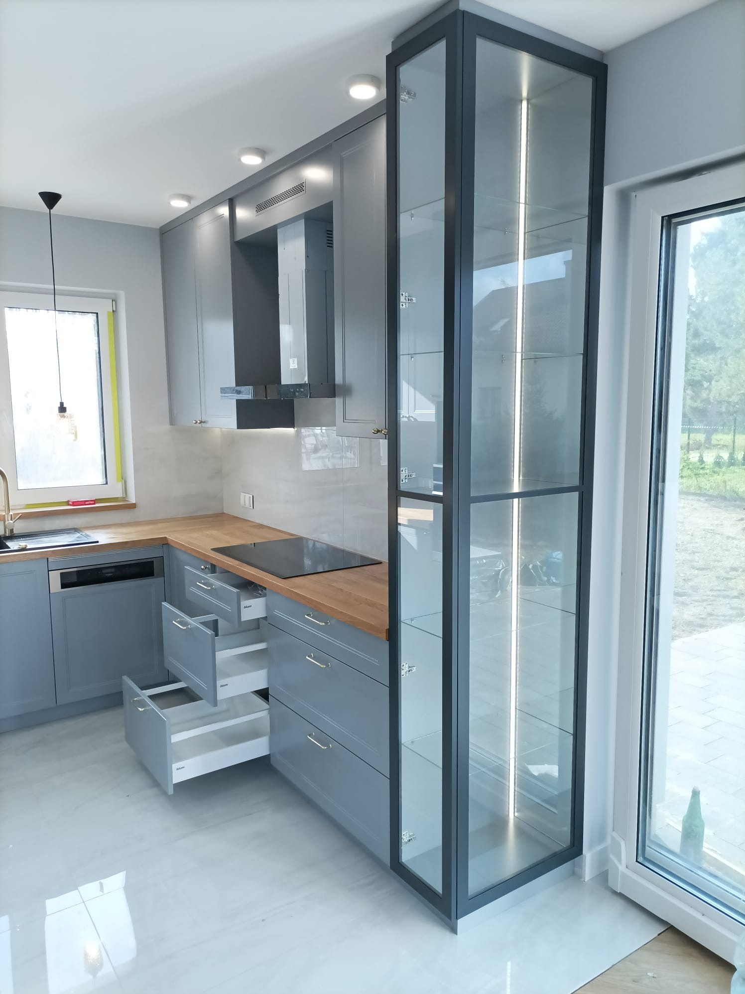 Szara kuchnia z drewnianym blatem i szklaną witryną z oświetleniem LED. Otwarta szuflada ukazuje system organizacji. Nowoczesny design, jasne wnętrze.