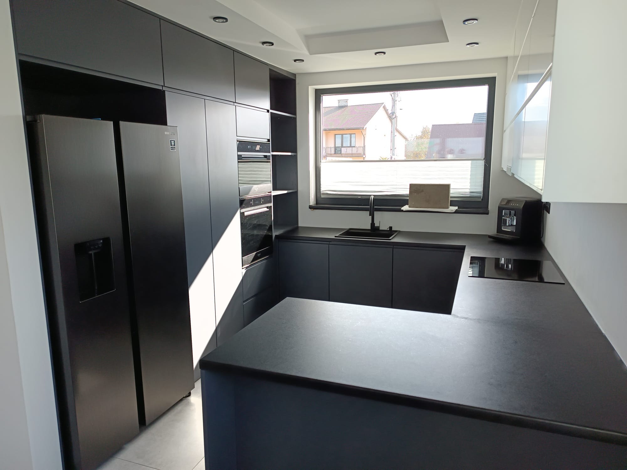 Nowoczesna, czarna kuchnia z matowymi frontami, dużym oknem i wyspą. Minimalistyczny design z zabudowaną lodówką i piekarnikiem. Widok na dom z ogrodem przez okno.