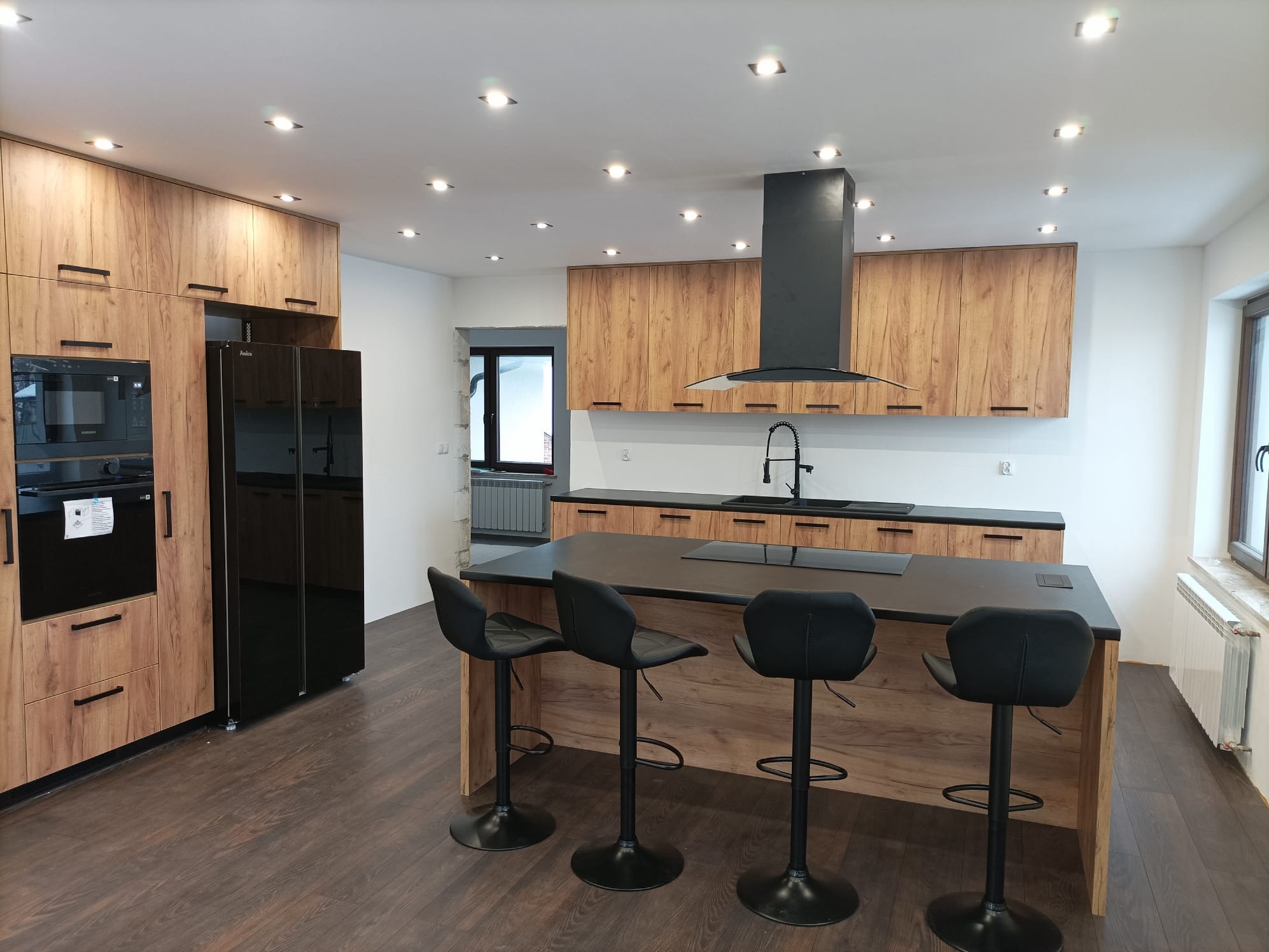 Nowoczesna kuchnia z drewnianymi frontami, czarnym blatem i wyspą z hokerami. Oświetlenie punktowe, czarny okap i sprzęt AGD. Ciemna podłoga.