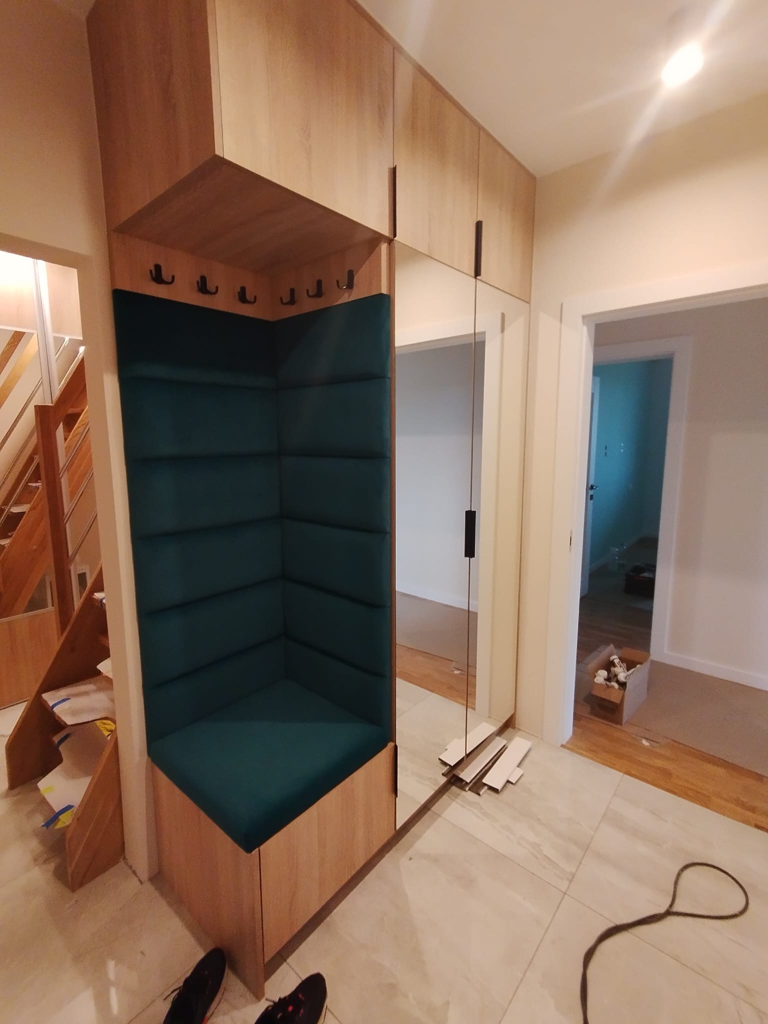 Funkcjonalna zabudowa przedpokoju z siedziskiem obitym zieloną tkaniną, wkomponowana w szafę z lustrem. Widoczne schody i fragment podłogi z butami.