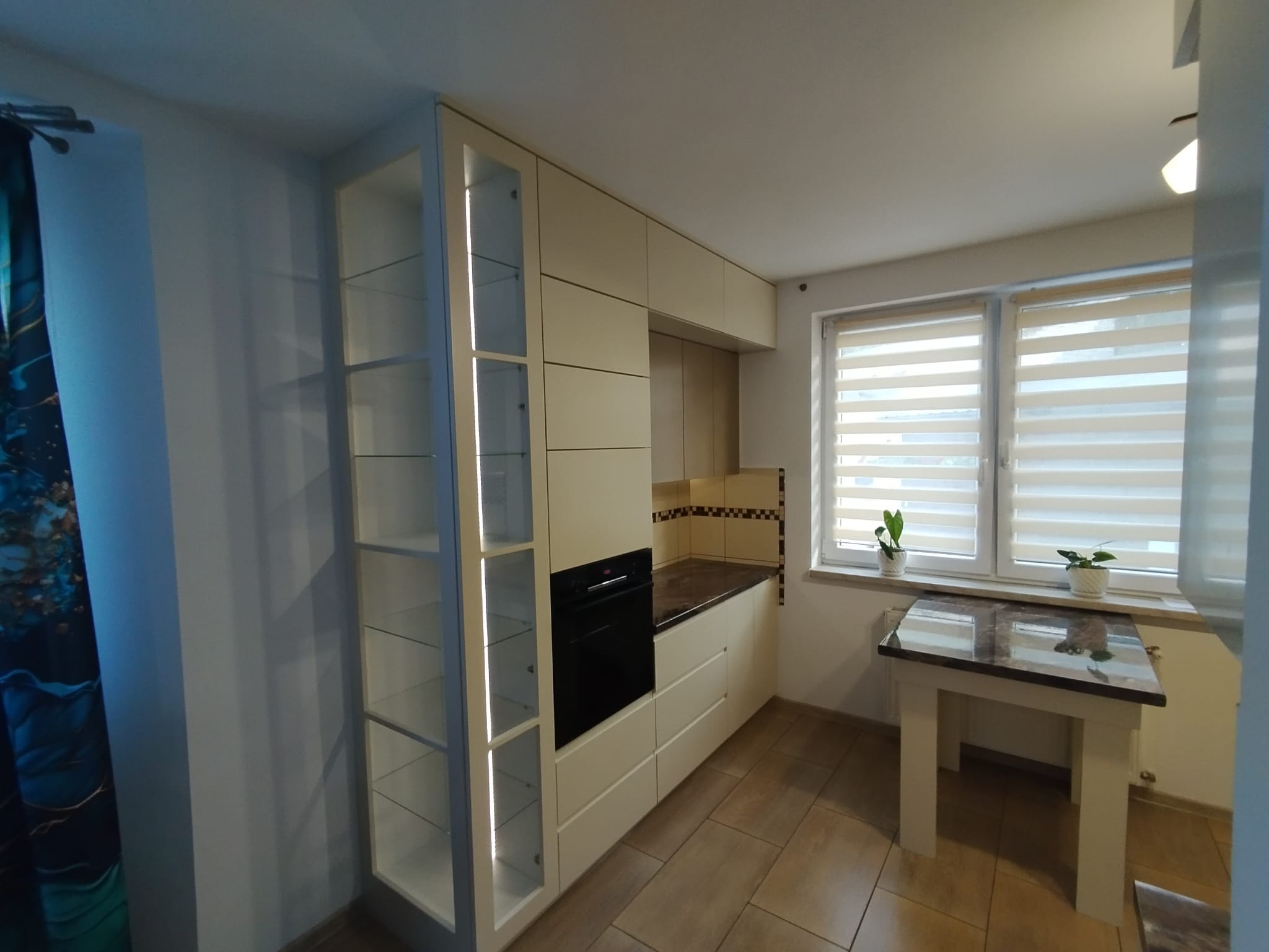 Jasna kuchnia z zabudową na wymiar: szafki wiszące, piekarnik, przeszklona witryna z oświetleniem LED. Blat z ciemnego kamienia, okno z roletami i stolik.