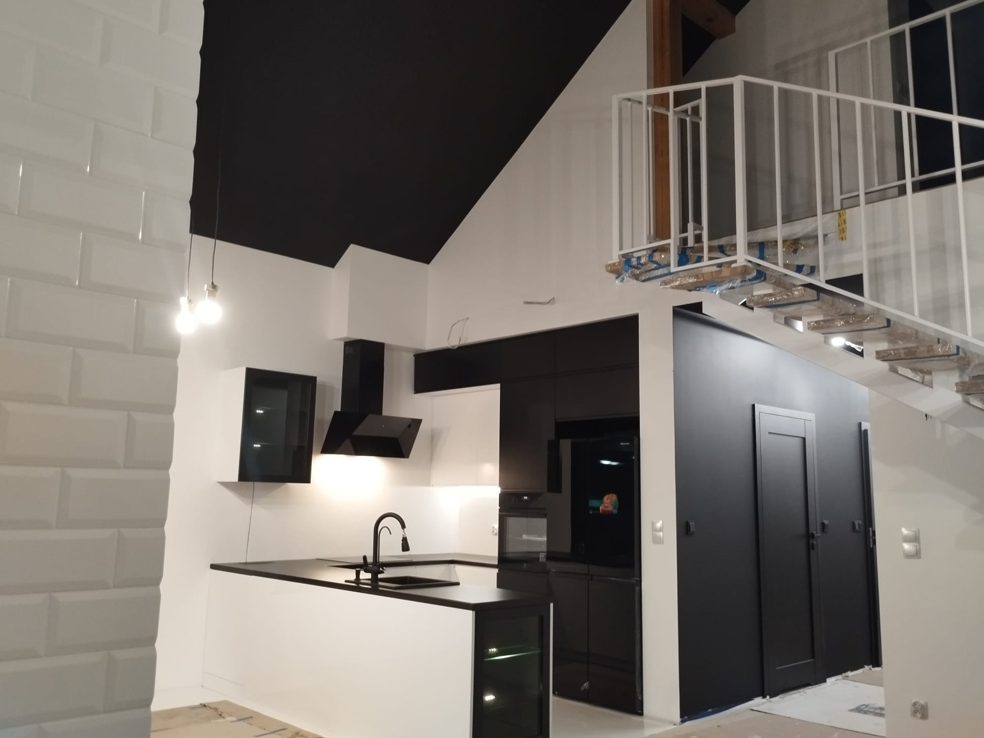 Nowoczesna kuchnia w czerni i bieli z wyspą, minimalistycznym okapem i schodami z metalową balustradą w tle. Kontrastowe połączenie kolorów i oświetlenie.