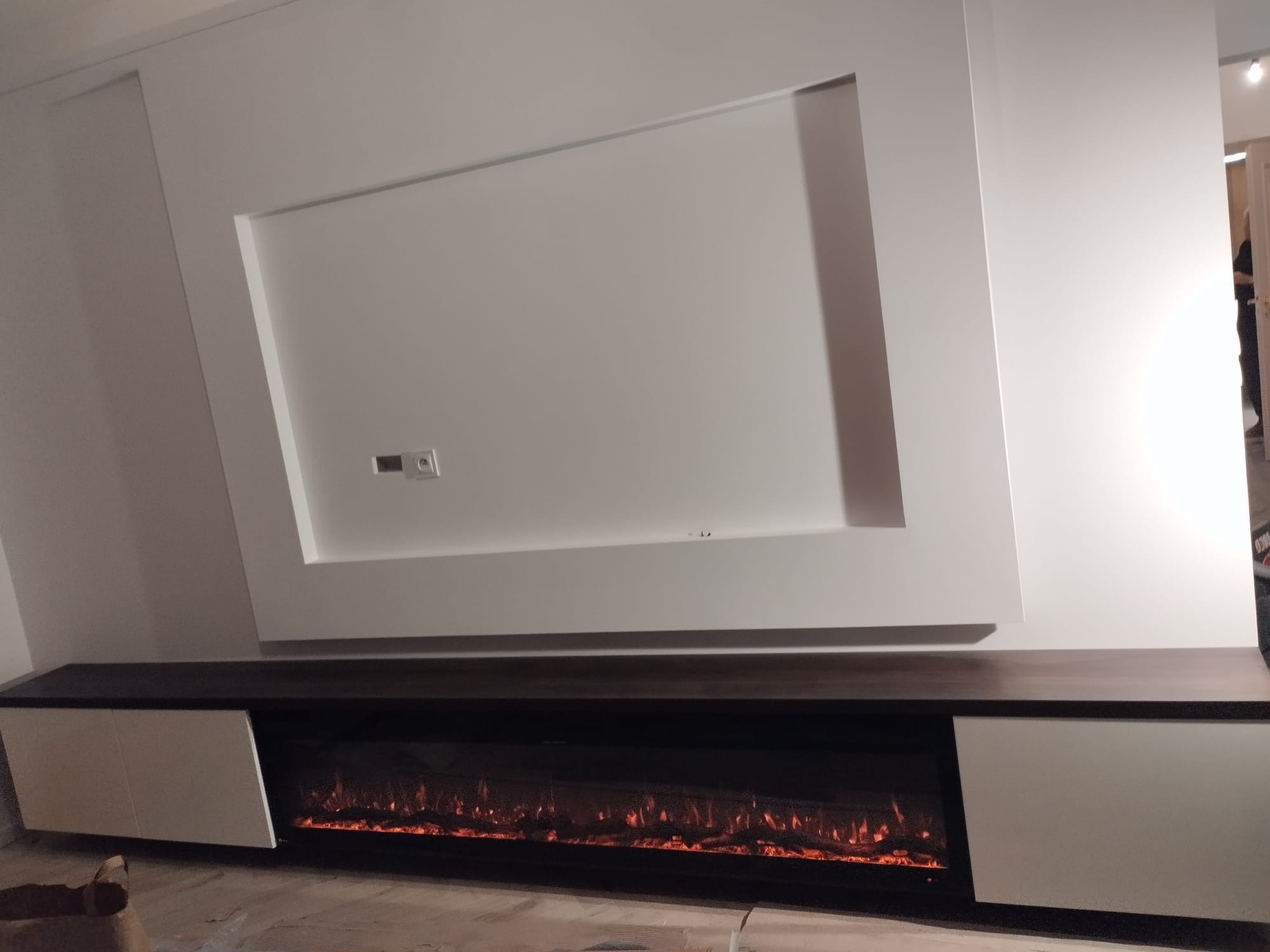 Nowoczesna szafka RTV z wbudowanym elektrycznym kominkiem i dekoracyjną ramą na ścianie. Minimalistyczny design, białe fronty szafek, ciemny blat i efekt płomieni.