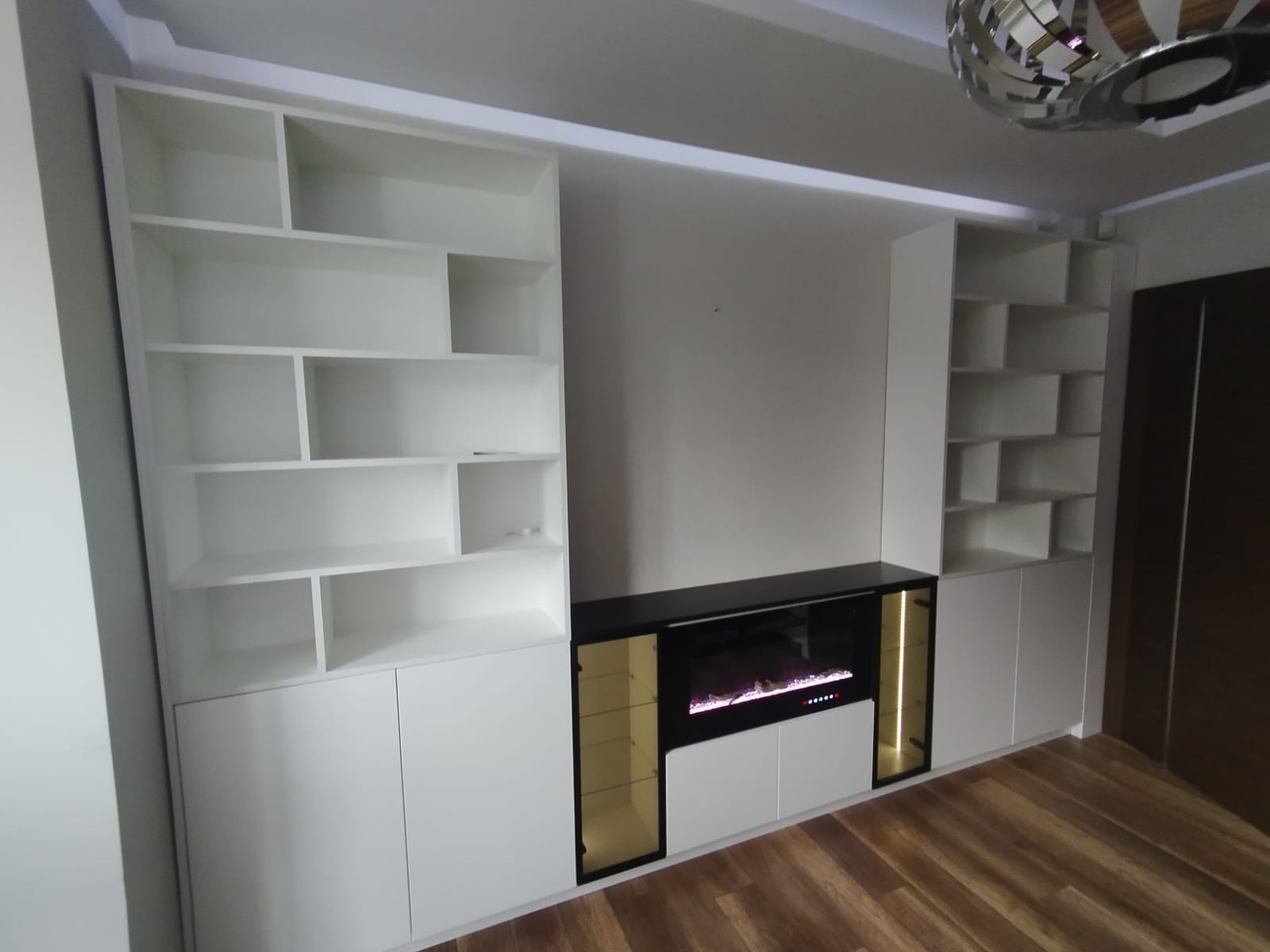 Biała zabudowa meblowa na ścianie z elektrycznym kominkiem i półkami. Nowoczesny design, minimalistyczny styl. Oświetlenie LED podkreśla detale.