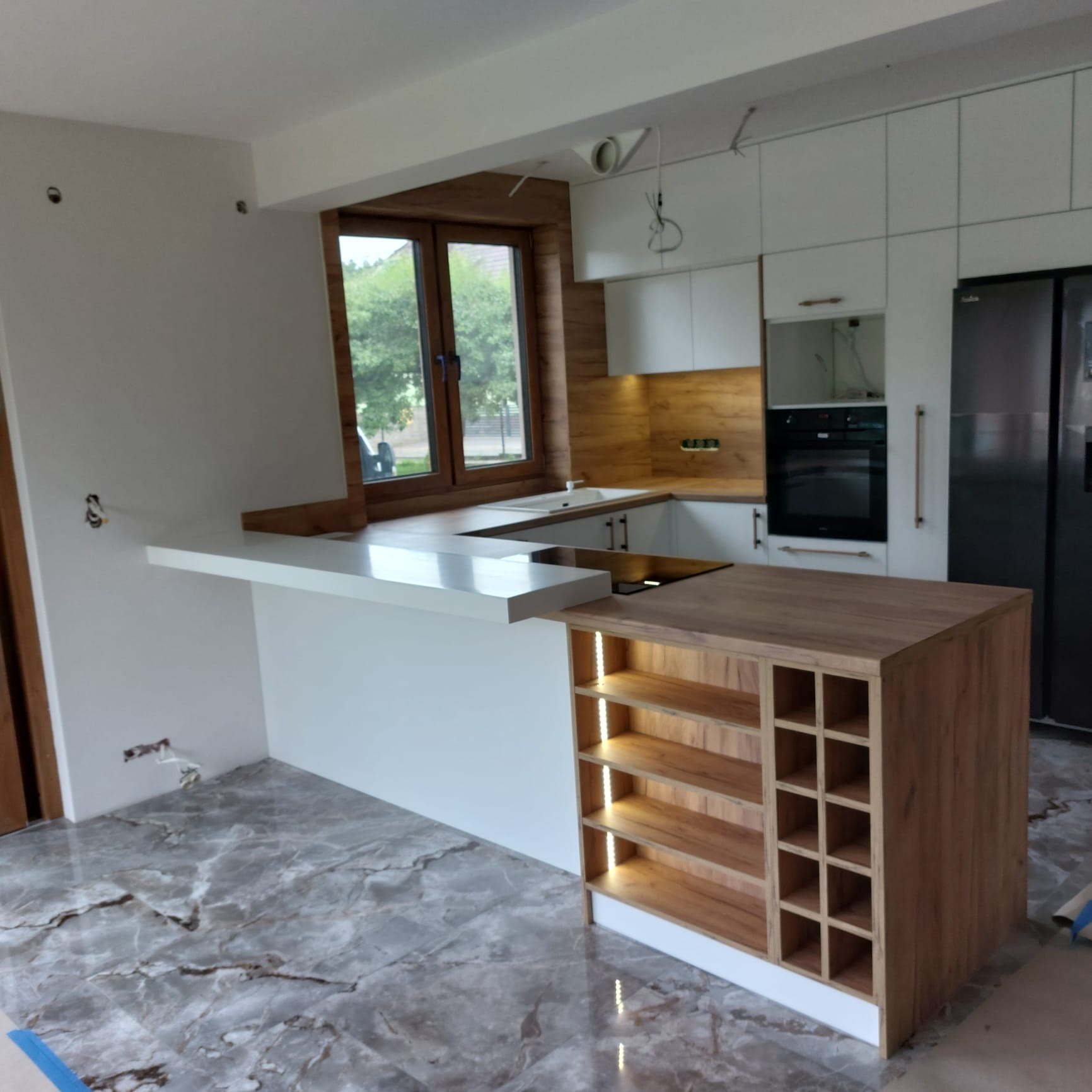 Nowoczesna kuchnia z wyspą i oświetleniem LED, białe fronty szafek, drewniane akcenty, marmurowa podłoga. Widok na okno z zielenią w tle. Częściowo widoczne instalacje.