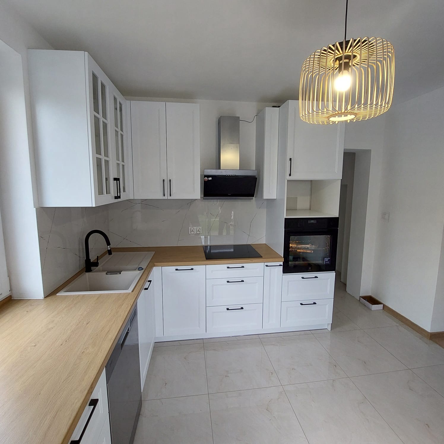 Jasna kuchnia z białymi szafkami i drewnianym blatem, czarne, minimalistyczne uchwyty, marmurowe wykończenie ściany, nowoczesna lampa wisząca.