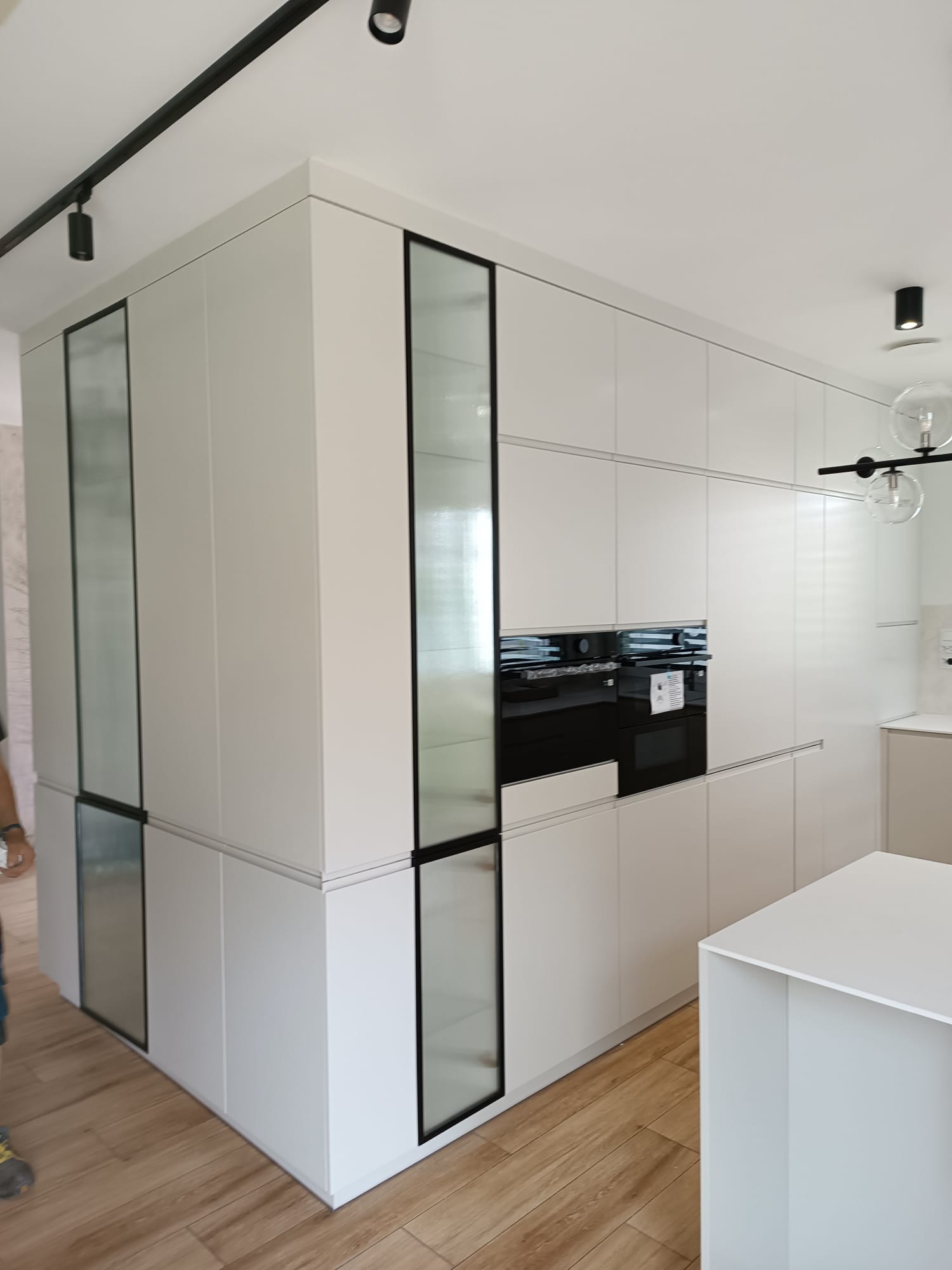 Nowoczesna, biała kuchnia na wymiar z czarnymi akcentami i szklanymi frontami w Modliborzycach. Minimalistyczny design, oświetlenie punktowe, drewniana podłoga.