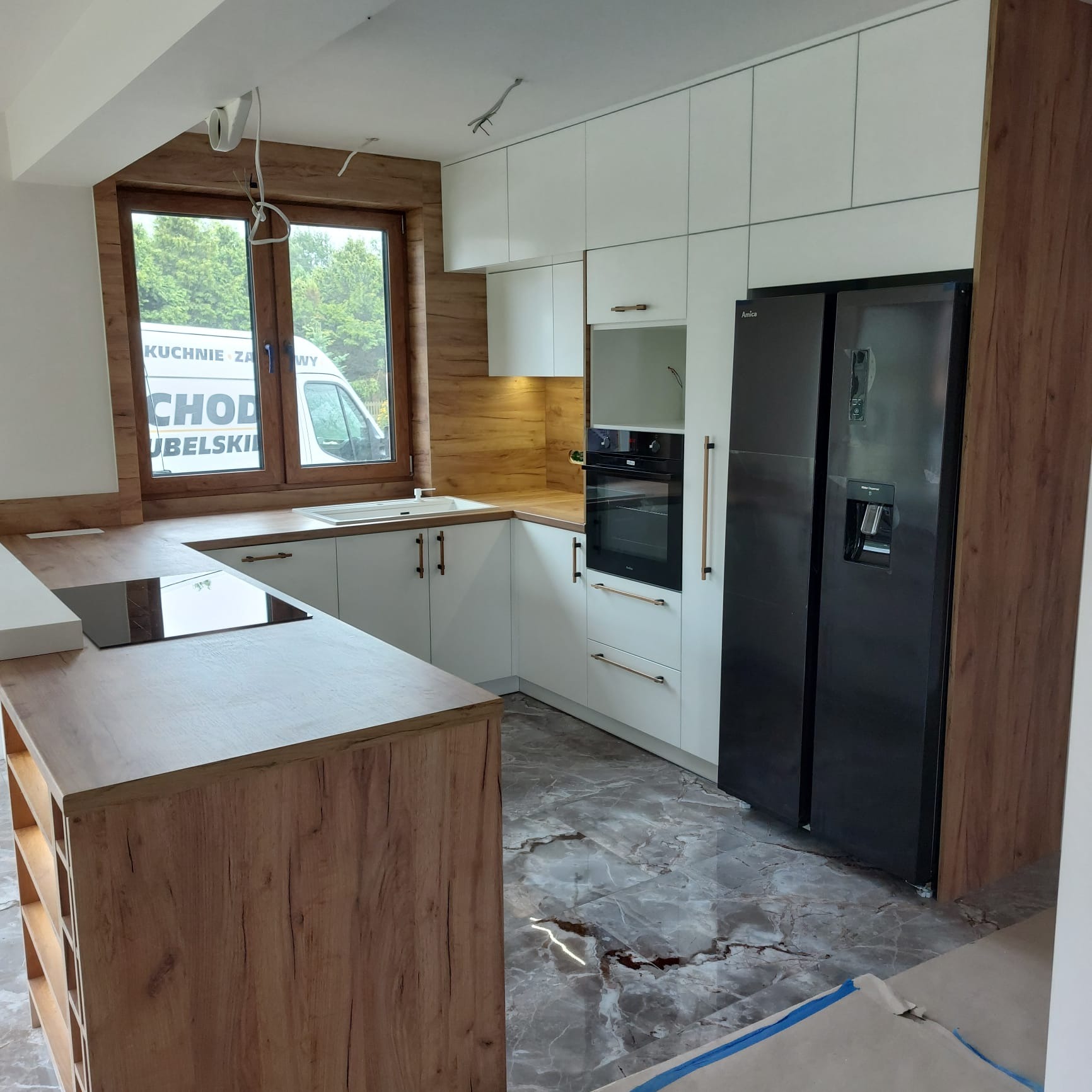 Nowoczesna kuchnia z białymi szafkami i drewnianymi akcentami, czarna lodówka Amica, marmurowe płytki podłogowe. Widok na okno z reklamą firmy.