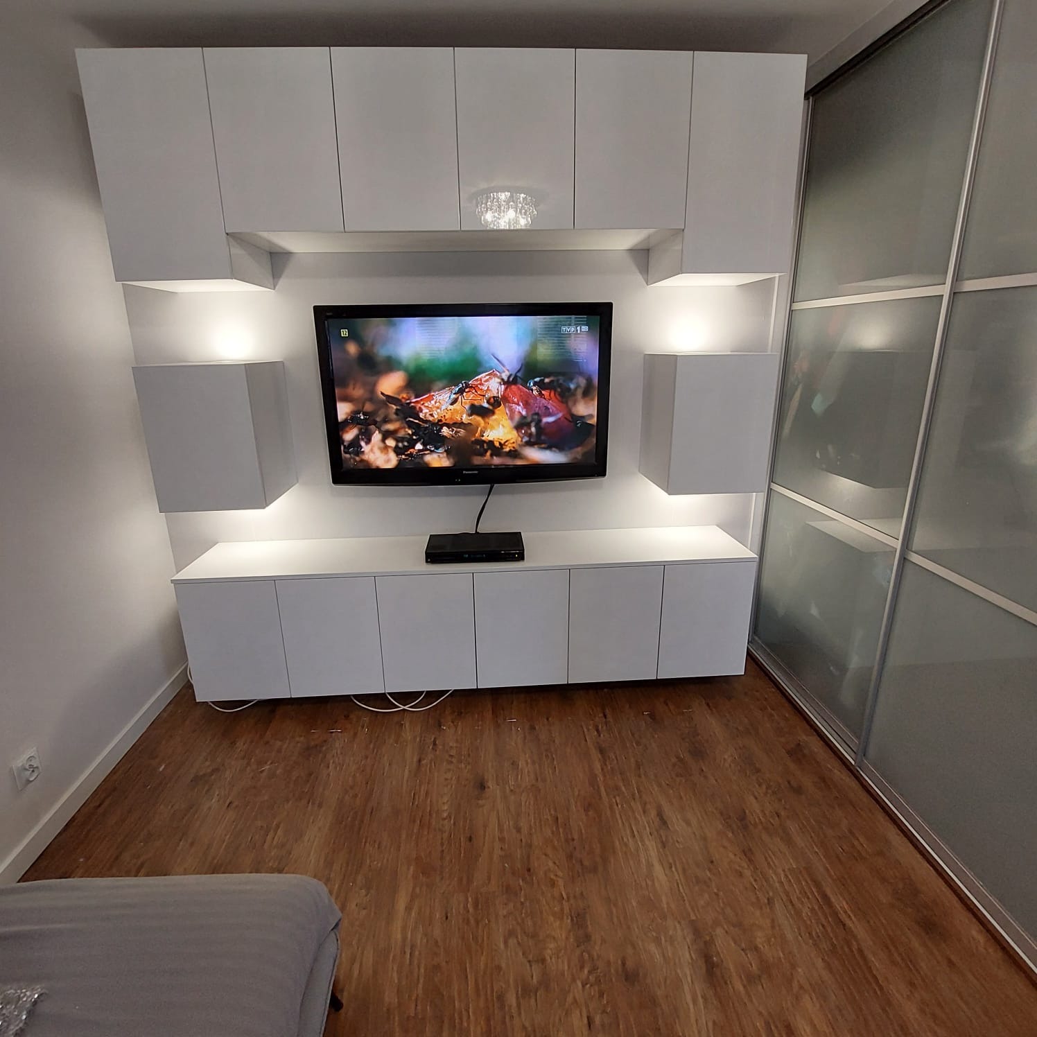 Biała zabudowa meblowa z szafkami, półkami i oświetleniem LED wokół telewizora, obok szafa przesuwna z matowego szkła, na podłodze panele imitujące drewno.