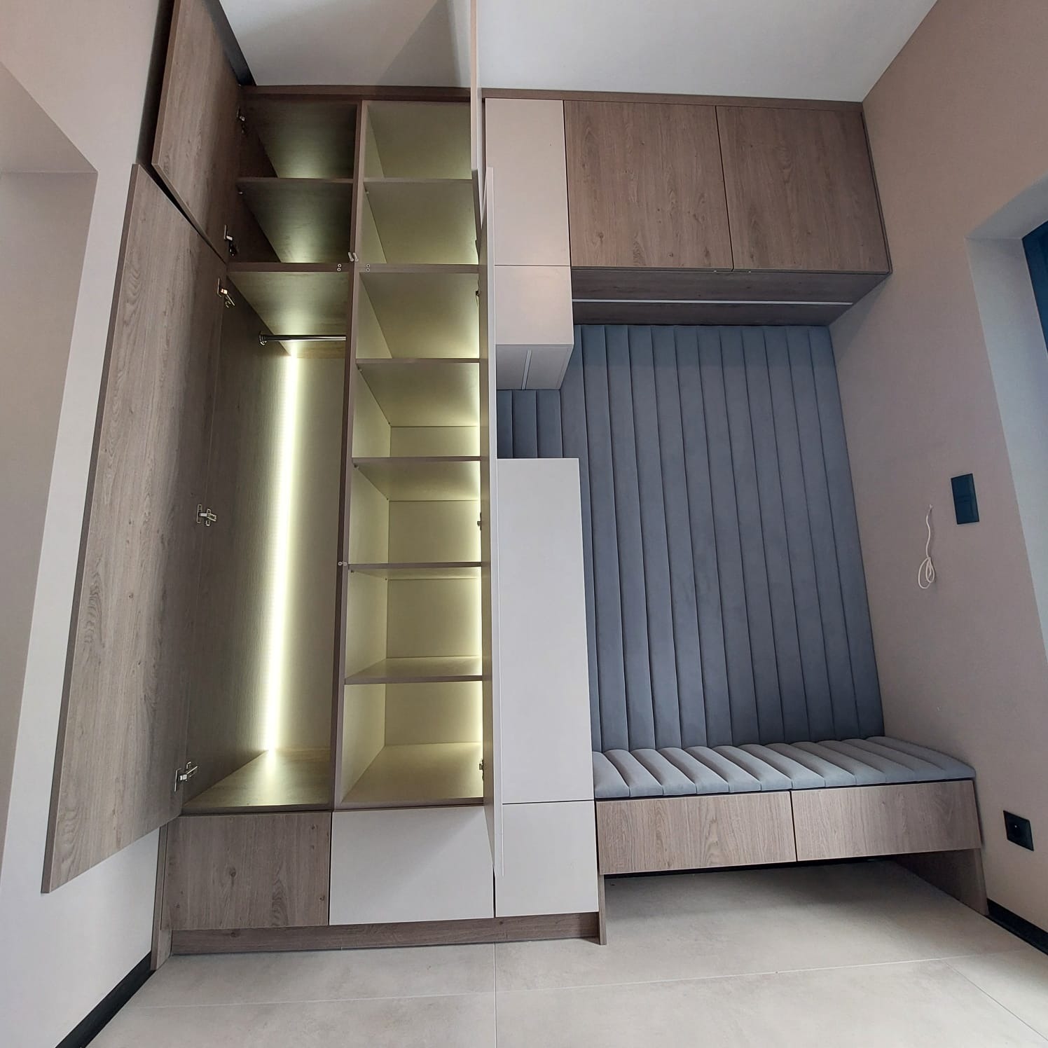 Funkcjonalna szafa wnękowa z oświetleniem LED i siedziskiem z tapicerowanym panelem ściennym w odcieniach szarości i drewna. Drzwiczki szafy otwarte, ukazują półki.