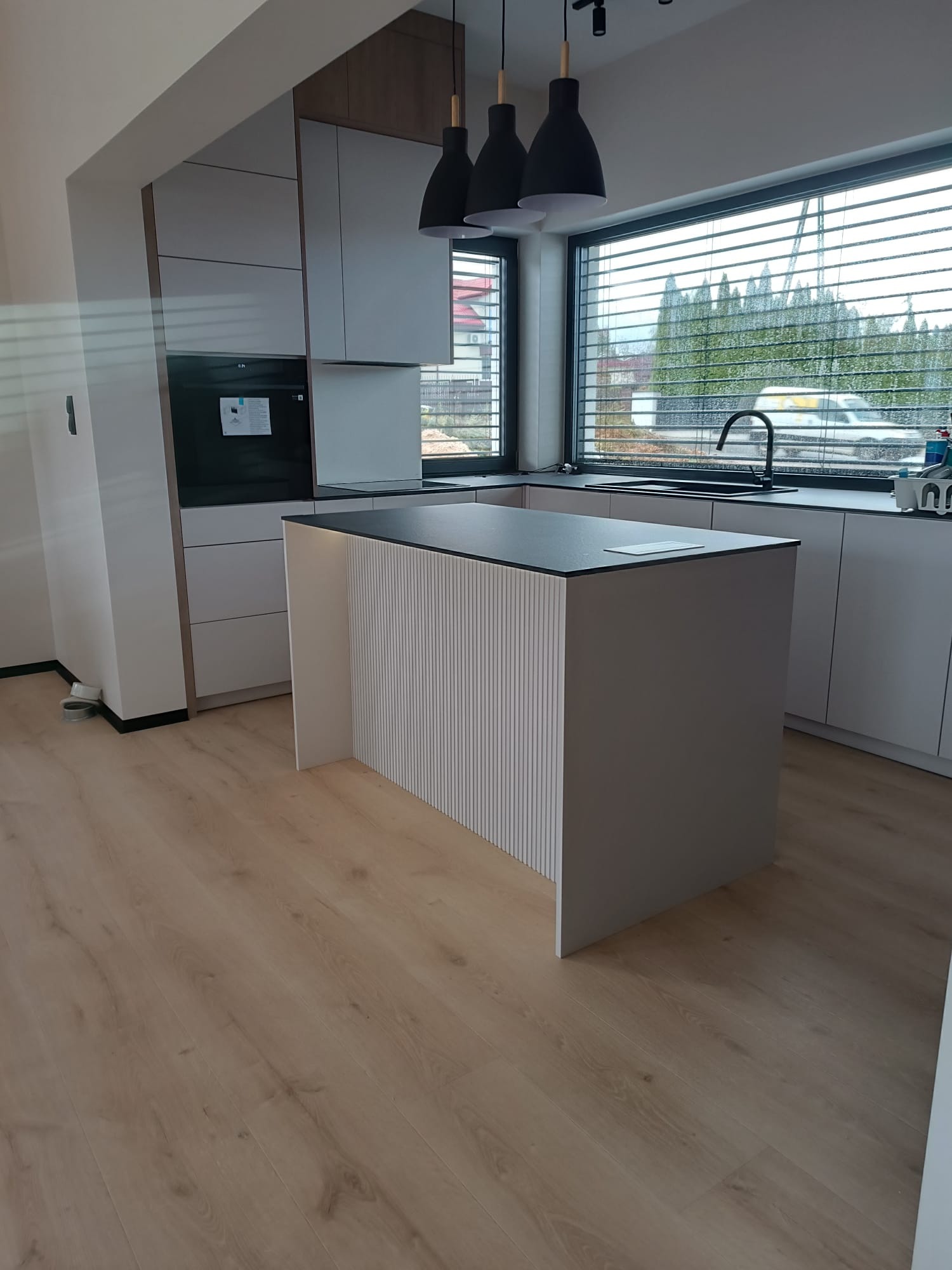 Nowoczesna kuchnia z wyspą i szarymi frontami, czarne lampy wiszące nad wyspą, duże okno z żaluzjami, jasna podłoga. Minimalistyczny design.