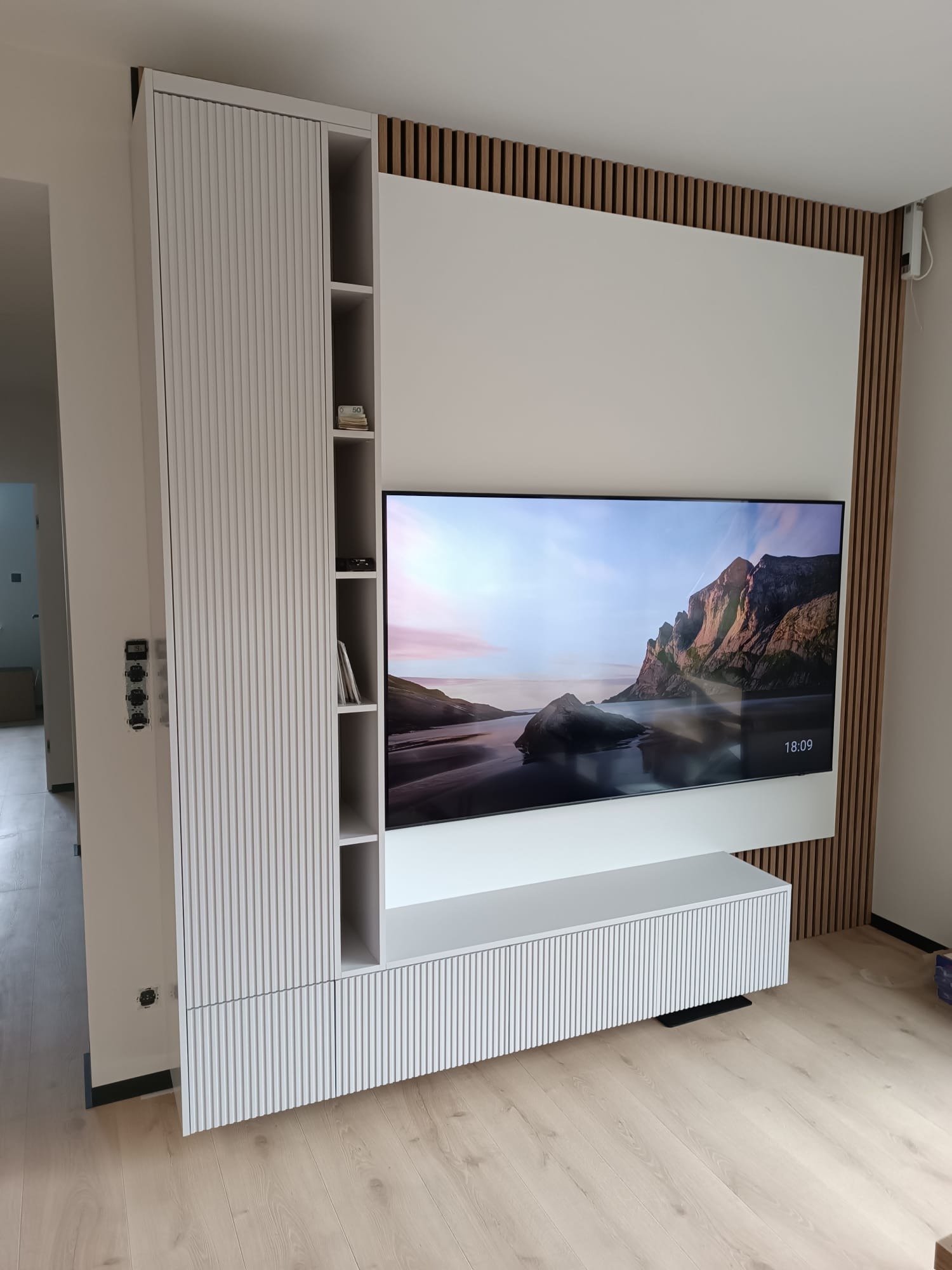 Biała zabudowa RTV z lamelami i szafką wiszącą, zintegrowana ze ścianą z drewnianymi listwami. Nowoczesny design, jasna podłoga, widok na inne pomieszczenie.