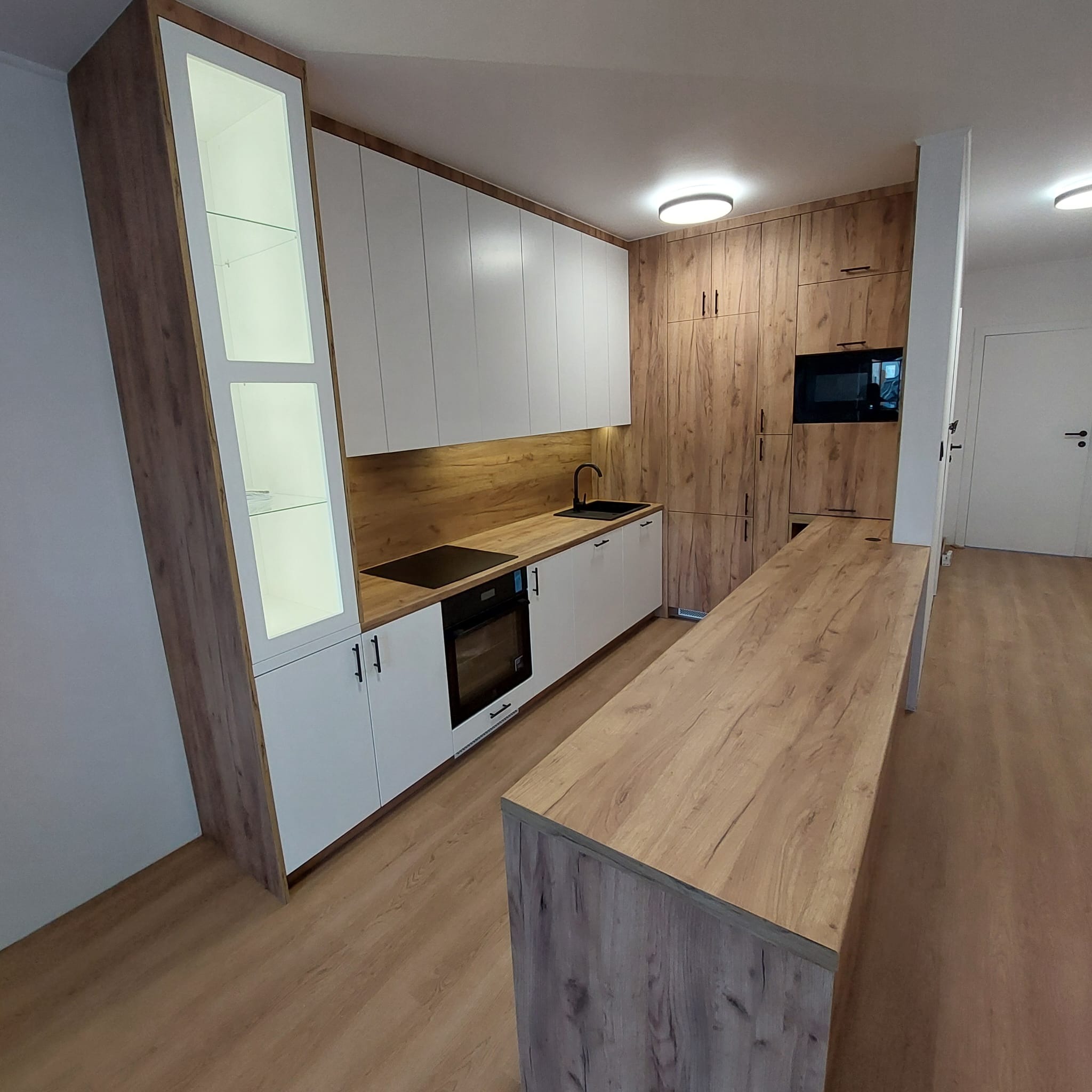 Nowoczesna kuchnia z białymi frontami i drewnianymi akcentami, oświetleniem LED i wyspą kuchenną z blatem w kolorze drewna. Widok na zabudowę kuchenną i sprzęt AGD.