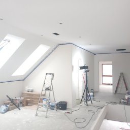 MK BUD - Białe poddasze po szpachlowaniu, zabezpieczone taśmą malarską, z oknami dachowymi i rozstawionymi narzędziami: drabina, lampa na statywie, wiadra z farbą.