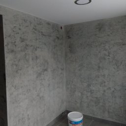 MK BUD - Pokój po dekoracyjnym malowaniu ścian w odcieniach szarości, z widocznym wiadrem farby Knauf Goldband Finish. Szare płytki podłogowe i okrągłe oświetlenie sufitowe.