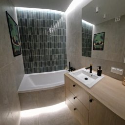 MK BUD - Nowoczesna łazienka z wanną, umywalką na drewnianej szafce i podświetleniem LED. Ciemne, pionowe płytki na ścianie dodają elegancji. Widoczne detale: czarna armatura i dekoracja.