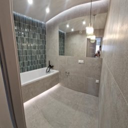 MK BUD - Nowoczesna łazienka w szarościach z akcentem zielonych płytek nad wanną. Lustro powiększa przestrzeń, a wiszące lampy dodają elegancji. Widoczny fragment osoby w odbiciu.