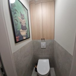 MK BUD - Wykończona toaleta z szarą glazurą, podwieszaną szafką z jasnego drewna i plakatem z kotem czytającym gazetę. Minimalistyczny design, czystość i funkcjonalność.