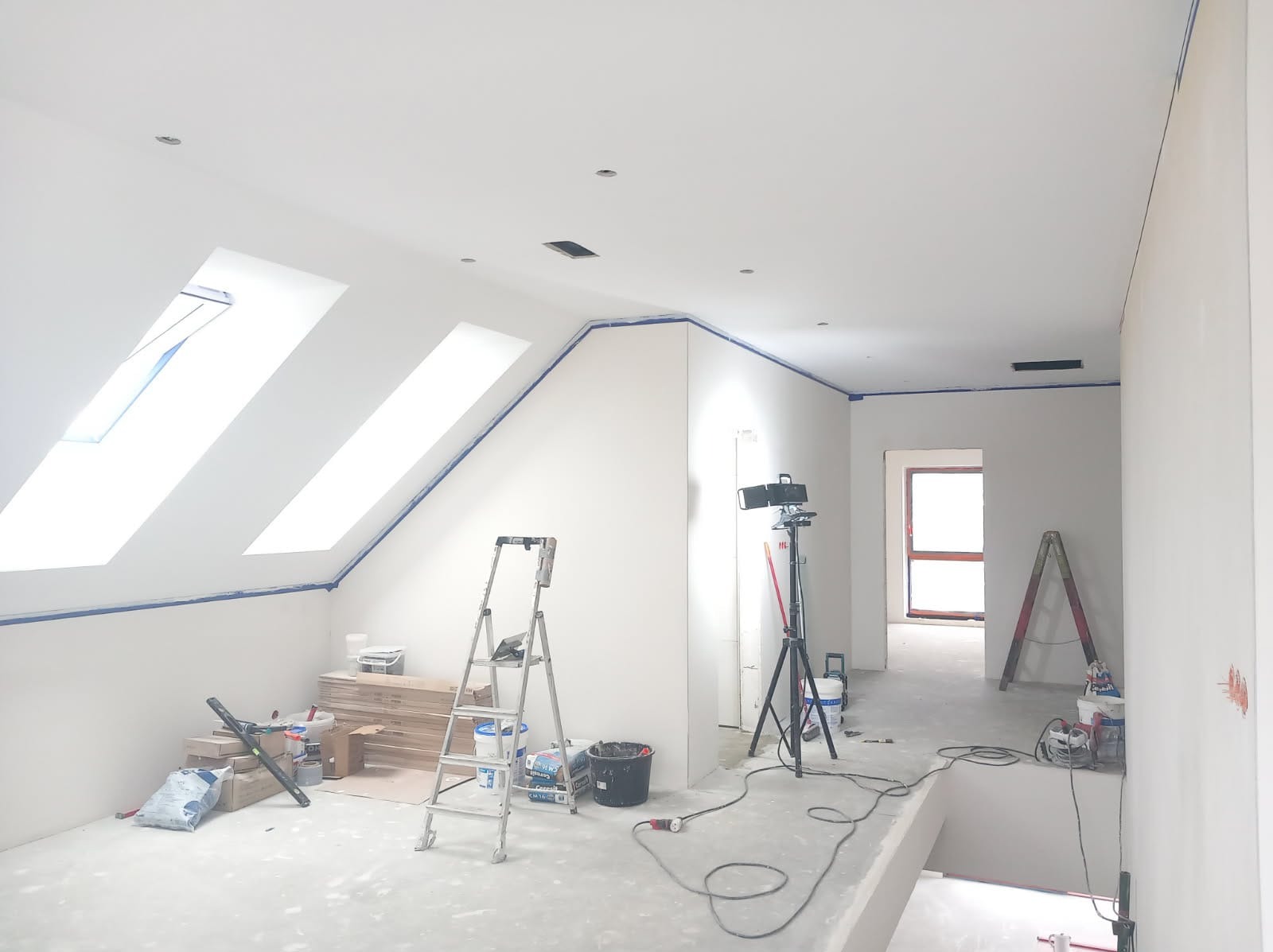 Białe poddasze po szpachlowaniu, zabezpieczone taśmą malarską, z oknami dachowymi i rozstawionymi narzędziami: drabina, lampa na statywie, wiadra z farbą.