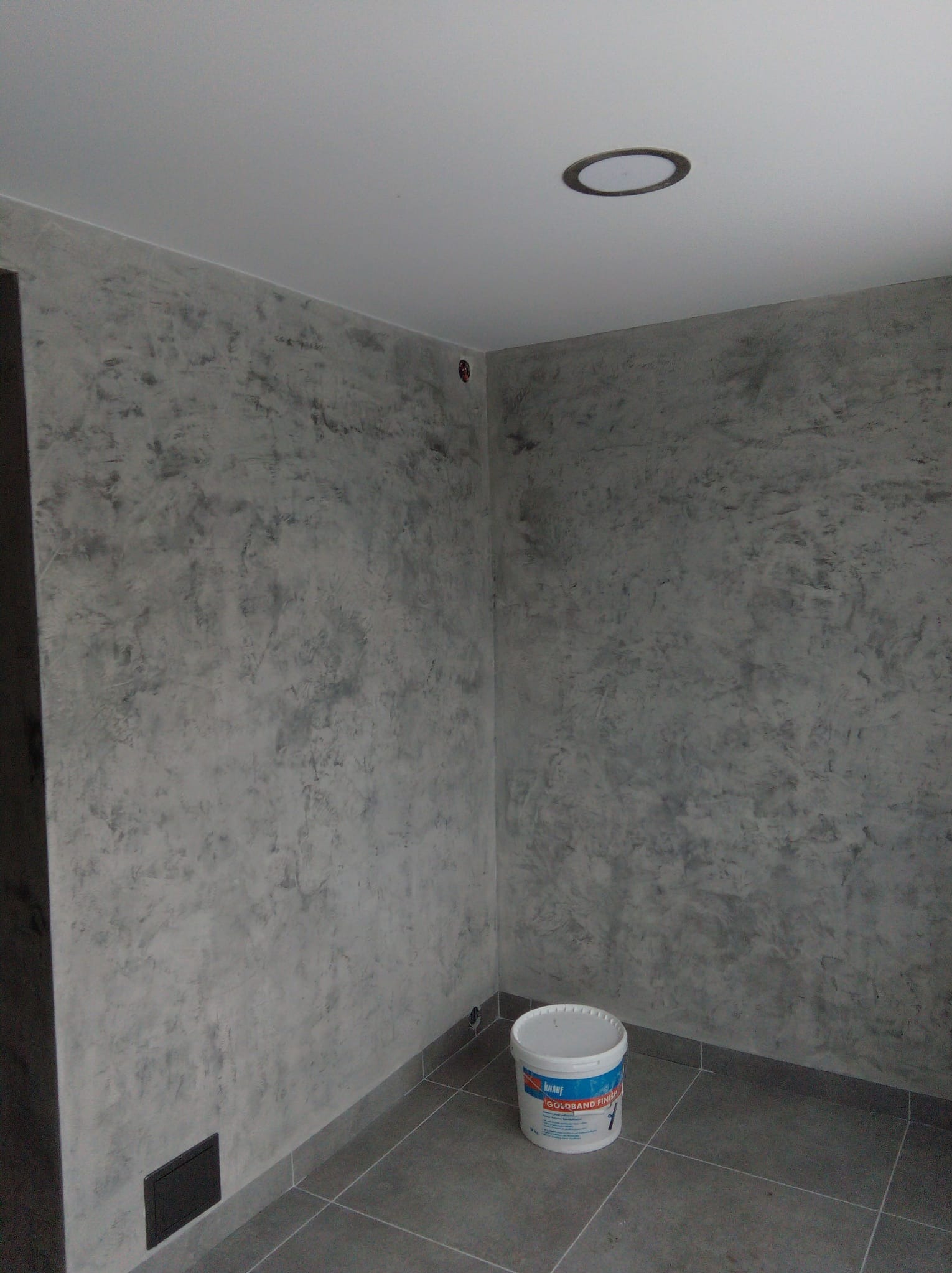 Pokój po dekoracyjnym malowaniu ścian w odcieniach szarości, z widocznym wiadrem farby Knauf Goldband Finish. Szare płytki podłogowe i okrągłe oświetlenie sufitowe.