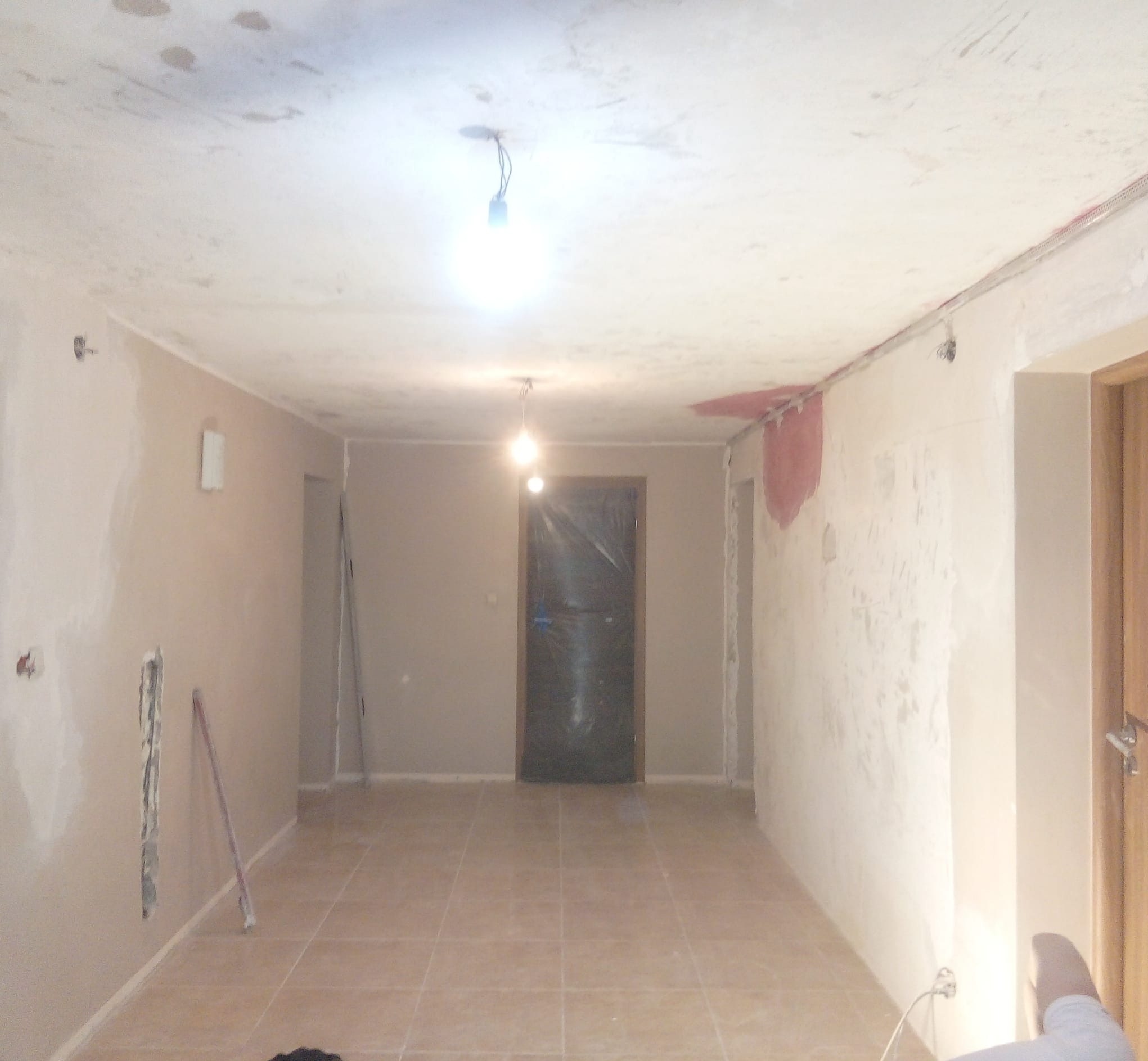 Wnętrze w trakcie remontu: ściany częściowo wyszpachlowane, widoczne instalacje elektryczne, podłoga wyłożona płytkami. Drzwi zabezpieczone folią.