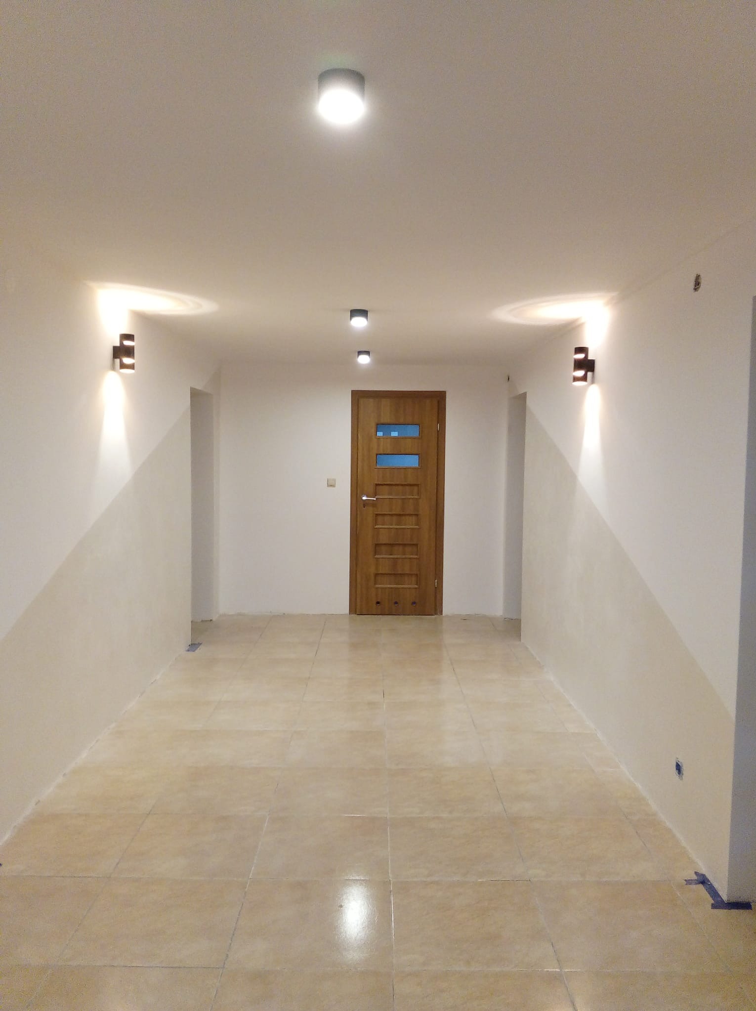 Wykończony korytarz z beżową podłogą, drewnianymi drzwiami z przeszkleniami i nowoczesnym oświetleniem ściennym. Ściany pomalowane w dwóch odcieniach bieli.