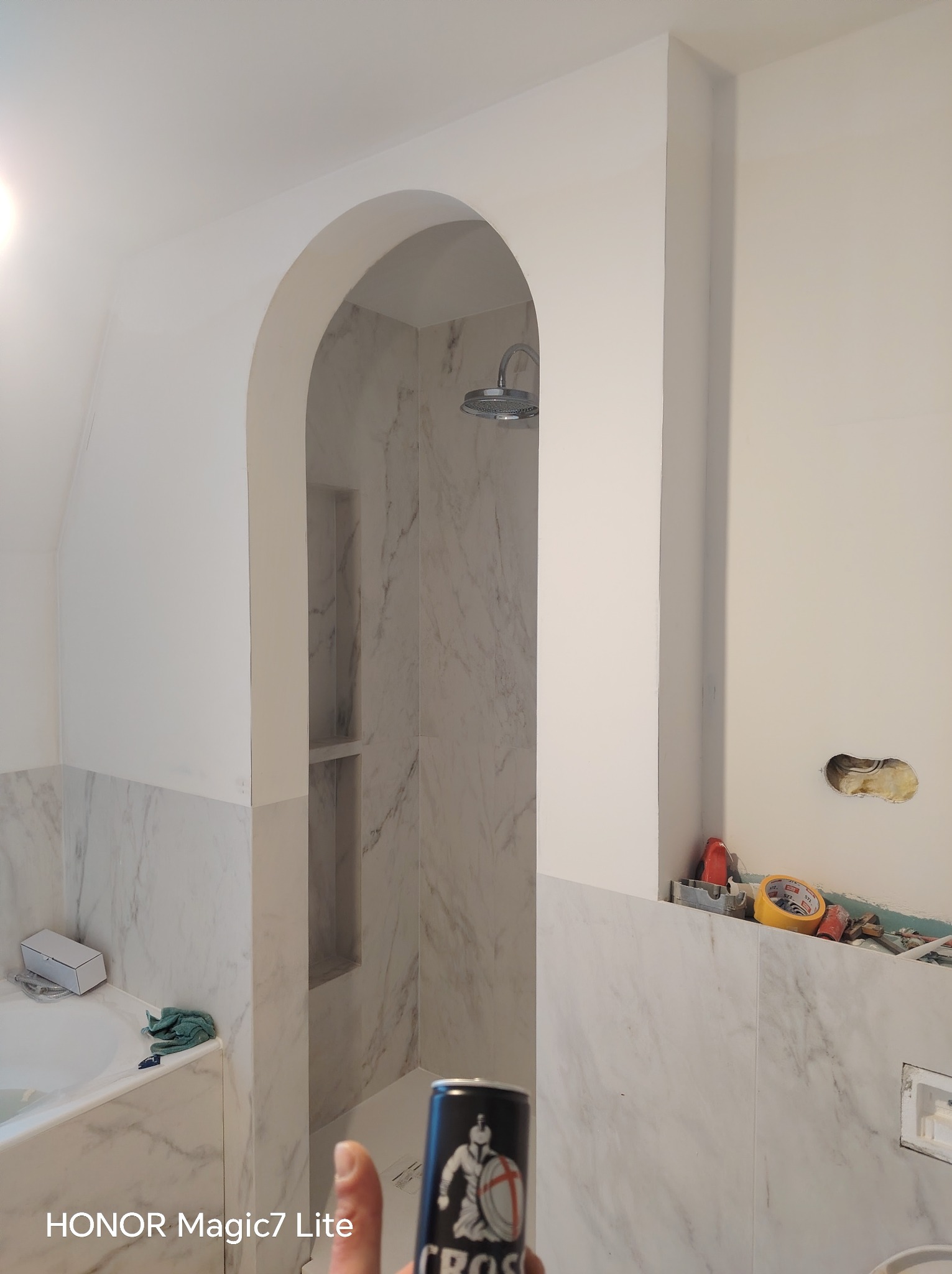 Łazienka w trakcie wykańczania: białe marmurowe płytki, kabina prysznicowa z wnękami, widoczne narzędzia i materiały budowlane. Otwór w ścianie przed wykończeniem.