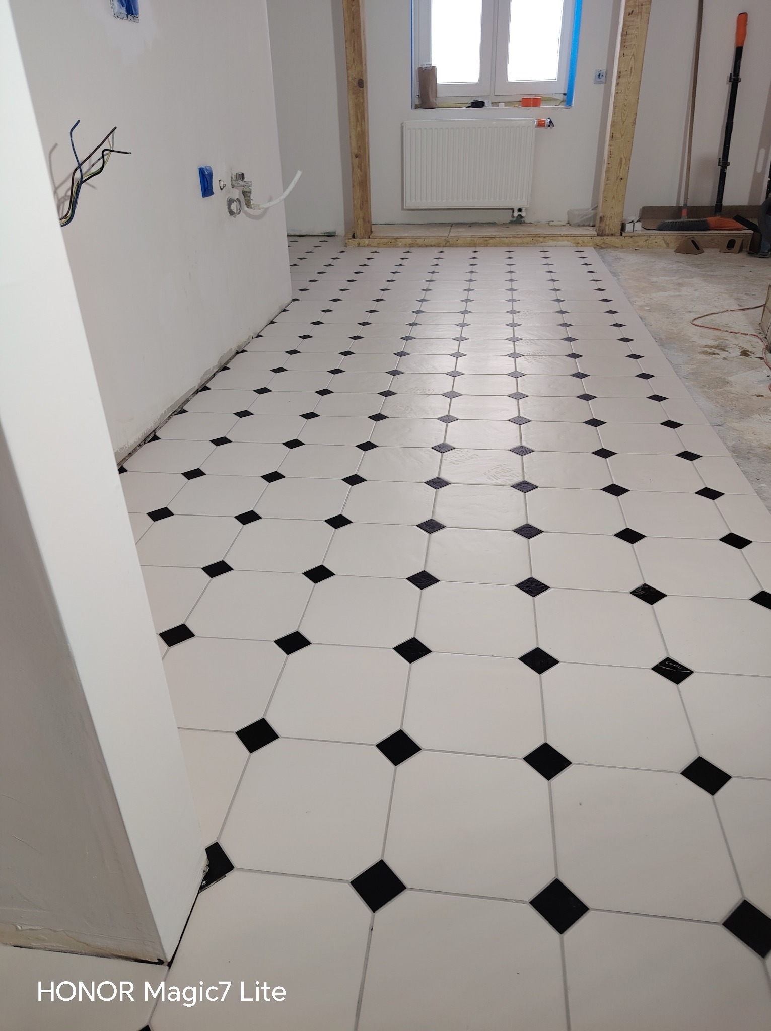 Ułożona podłoga z białych płytek z czarnymi wstawkami w romby, widok z góry, w tle instalacja elektryczna i grzejnik. Klasyczny wzór z precyzyjnym spasowaniem.