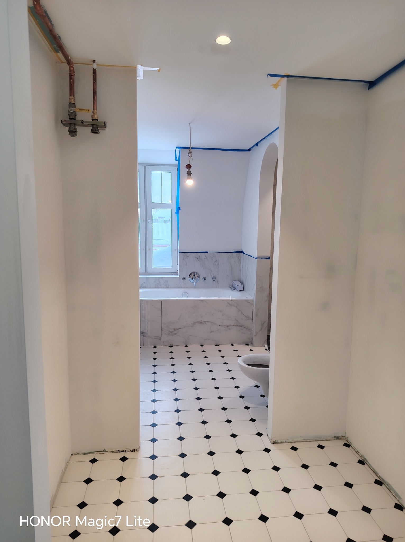 Białe ściany łazienki w trakcie malowania, zabezpieczone niebieską taśmą malarską. Widoczna armatura i fragment podłogi w biało-czarną szachownicę.