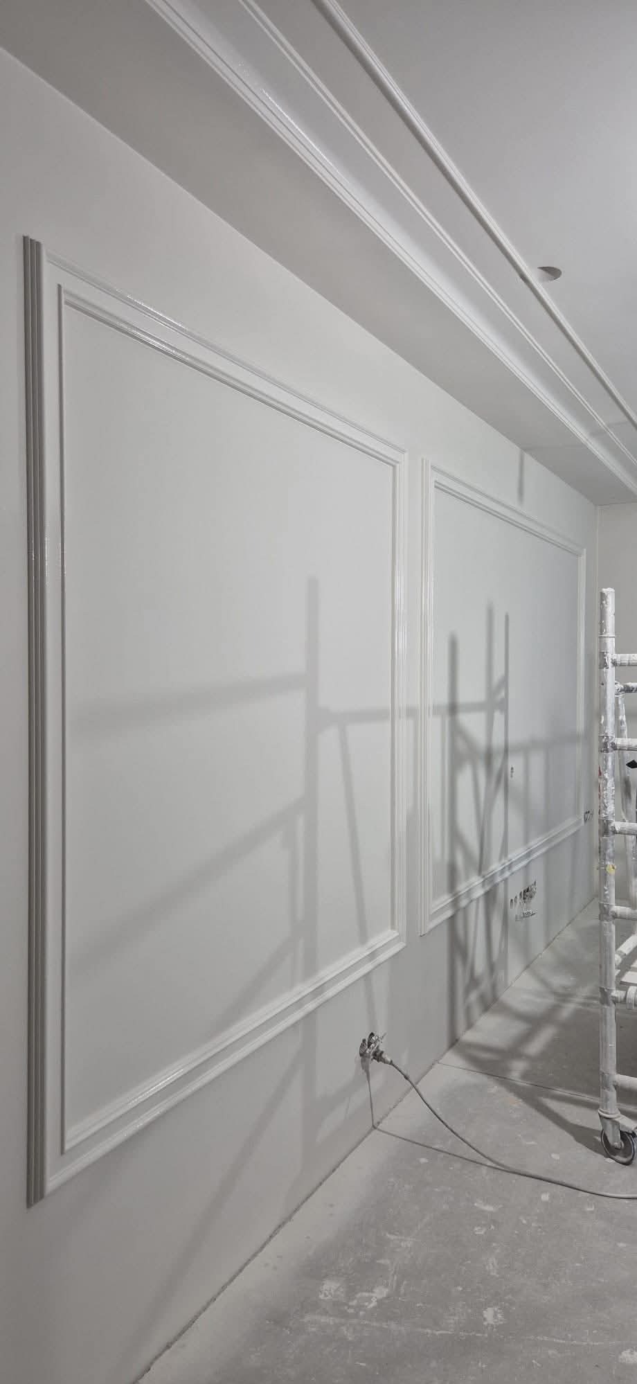 Świeżo pomalowana, biała ściana z dekoracyjnymi listwami i sztukaterią. Widoczny fragment rusztowania i przewód od urządzenia malarskiego.