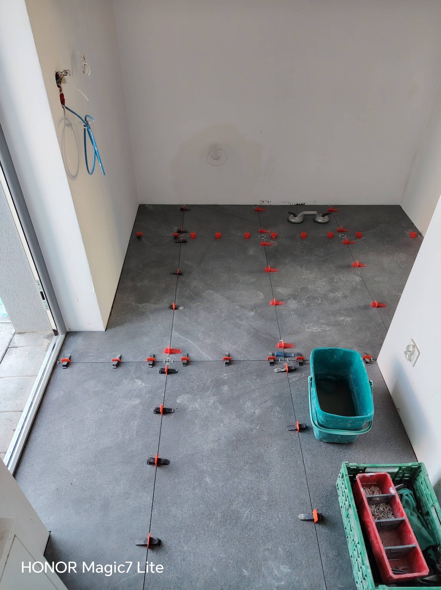 Układanie szarych płytek podłogowych z użyciem systemu poziomowania. Widoczne narzędzia glazurnicze: przyssawki, wiadro, skrzynka z akcesoriami. Świeżo położona podłoga w jasnym pomieszczeniu.