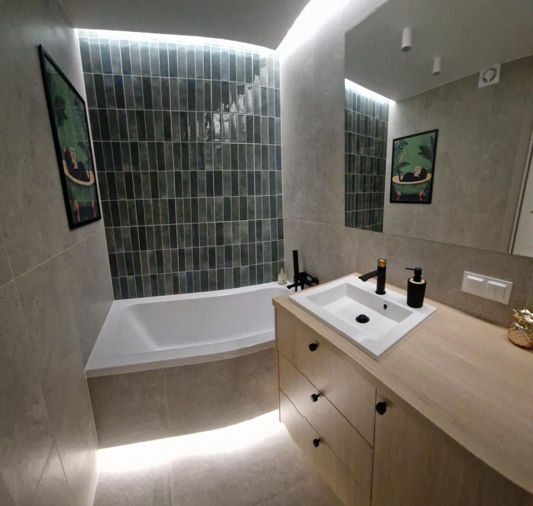 Nowoczesna łazienka z wanną, umywalką na drewnianej szafce i podświetleniem LED. Ciemne, pionowe płytki na ścianie dodają elegancji. Widoczne detale: czarna armatura i dekoracja.