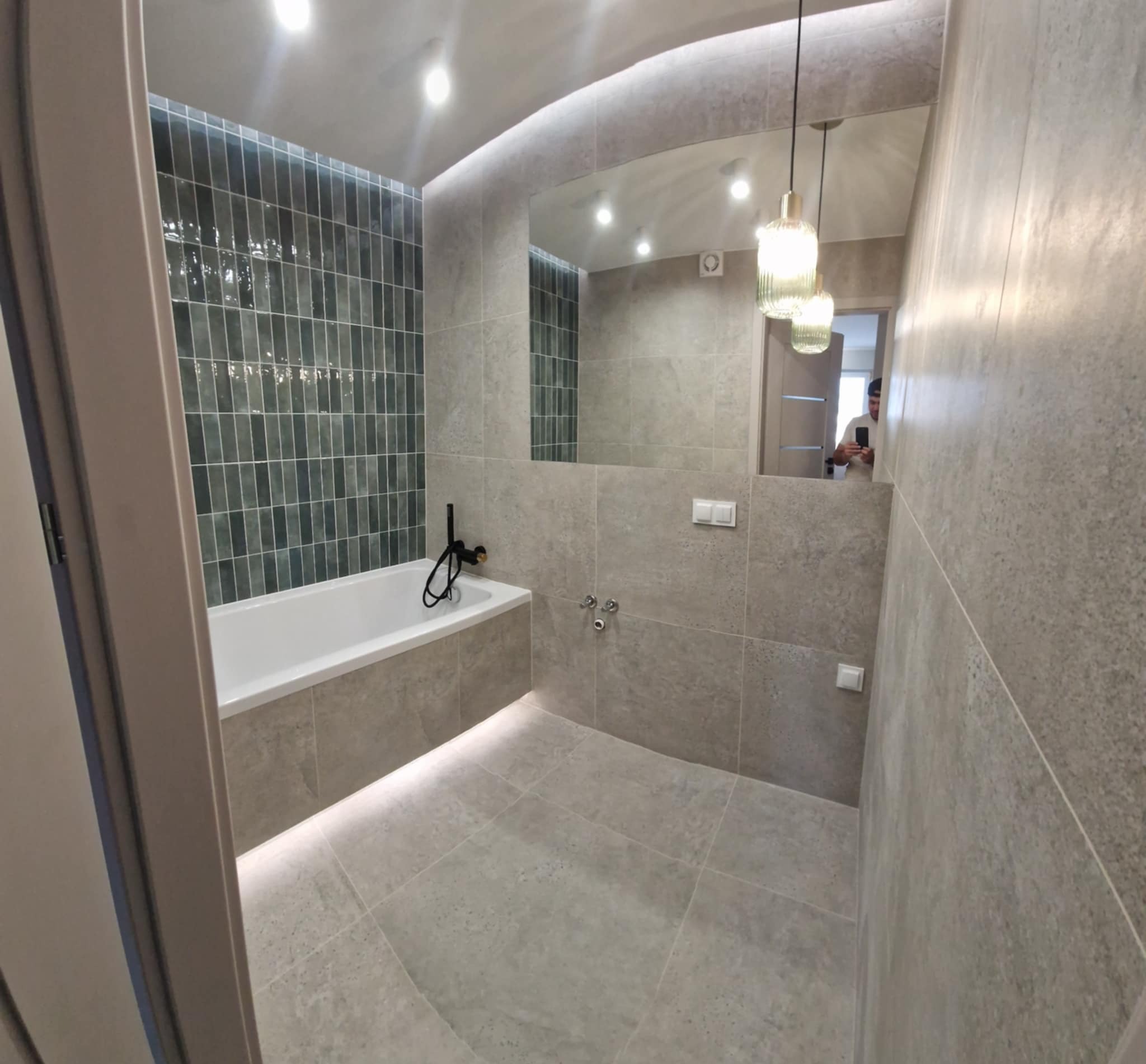 Nowoczesna łazienka w szarościach z akcentem zielonych płytek nad wanną. Lustro powiększa przestrzeń, a wiszące lampy dodają elegancji. Widoczny fragment osoby w odbiciu.