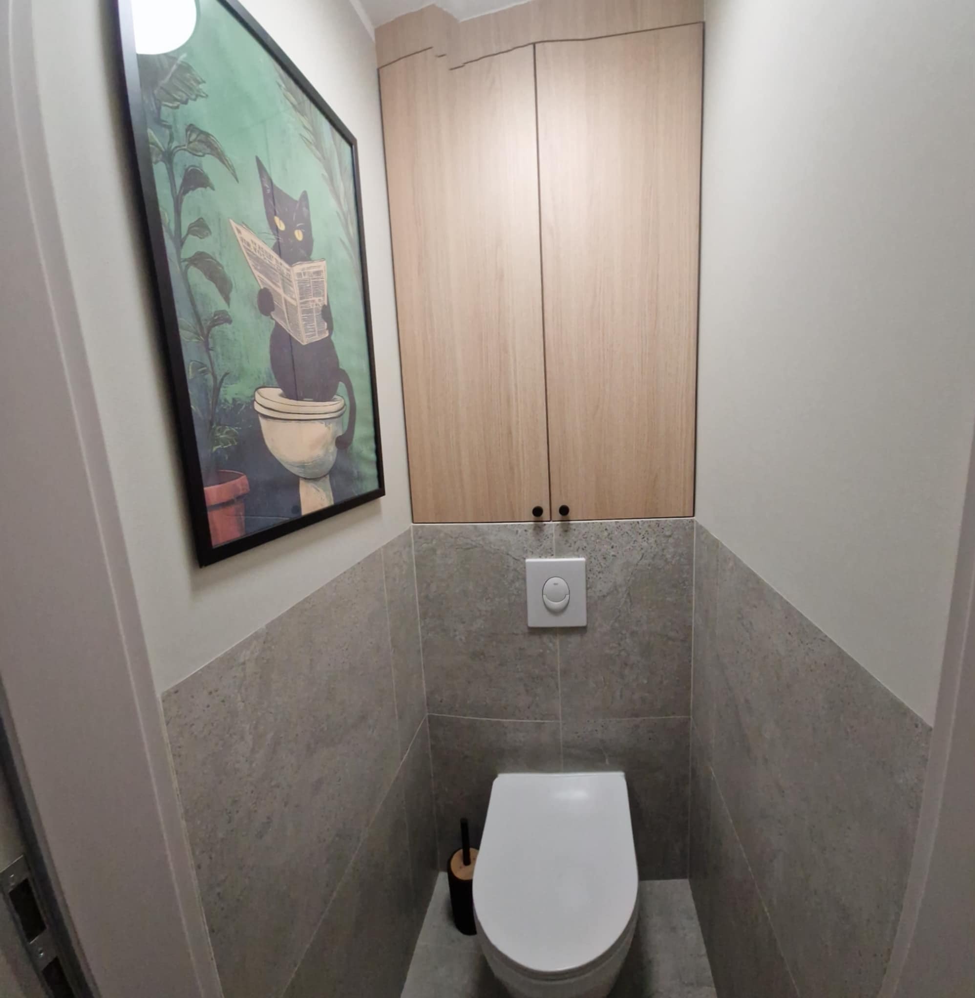 Wykończona toaleta z szarą glazurą, podwieszaną szafką z jasnego drewna i plakatem z kotem czytającym gazetę. Minimalistyczny design, czystość i funkcjonalność.