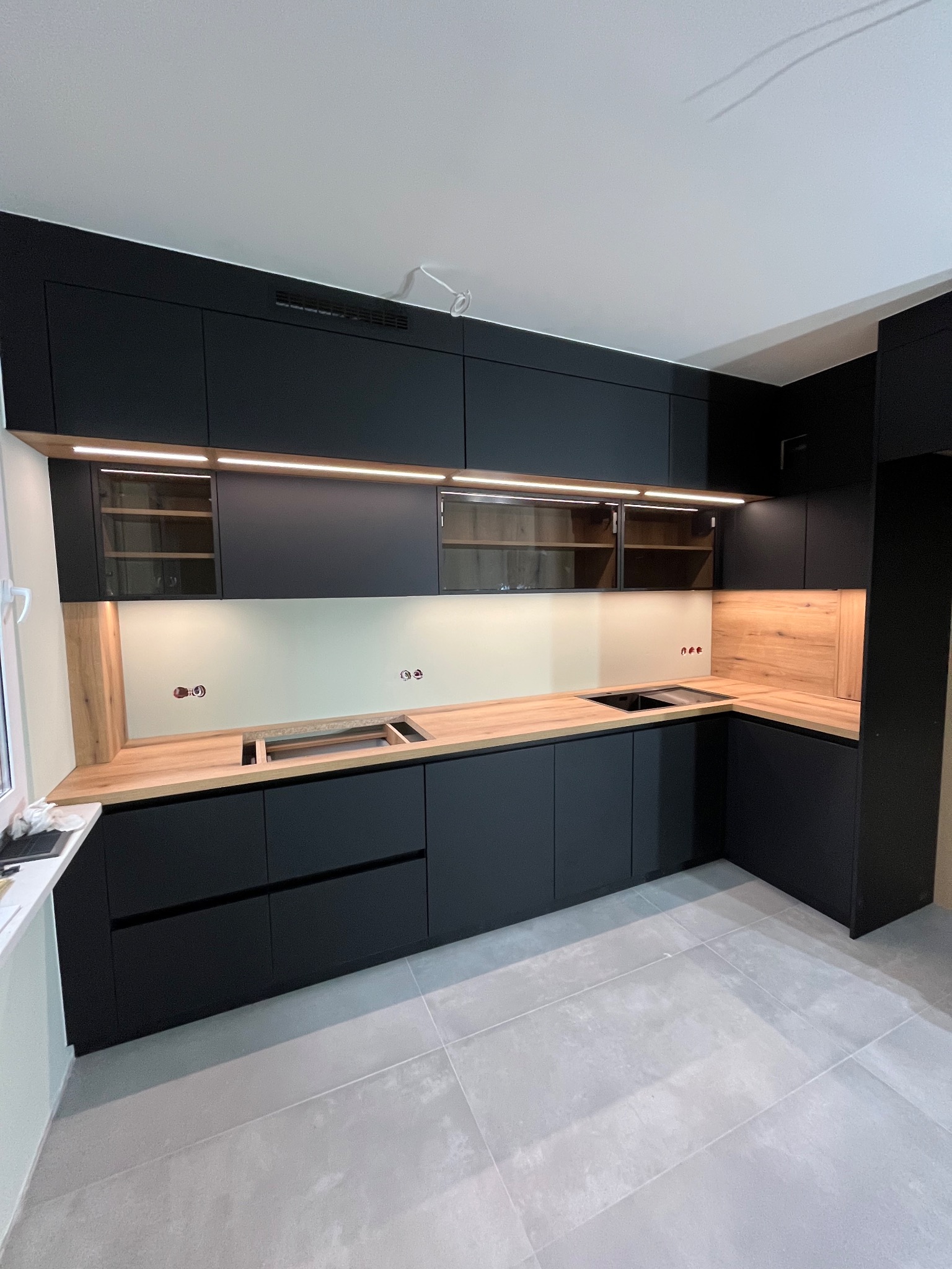 Nowoczesna, czarna zabudowa kuchenna z drewnianym blatem i oświetleniem LED, widok na narożną część kuchni z płytkami podłogowymi.