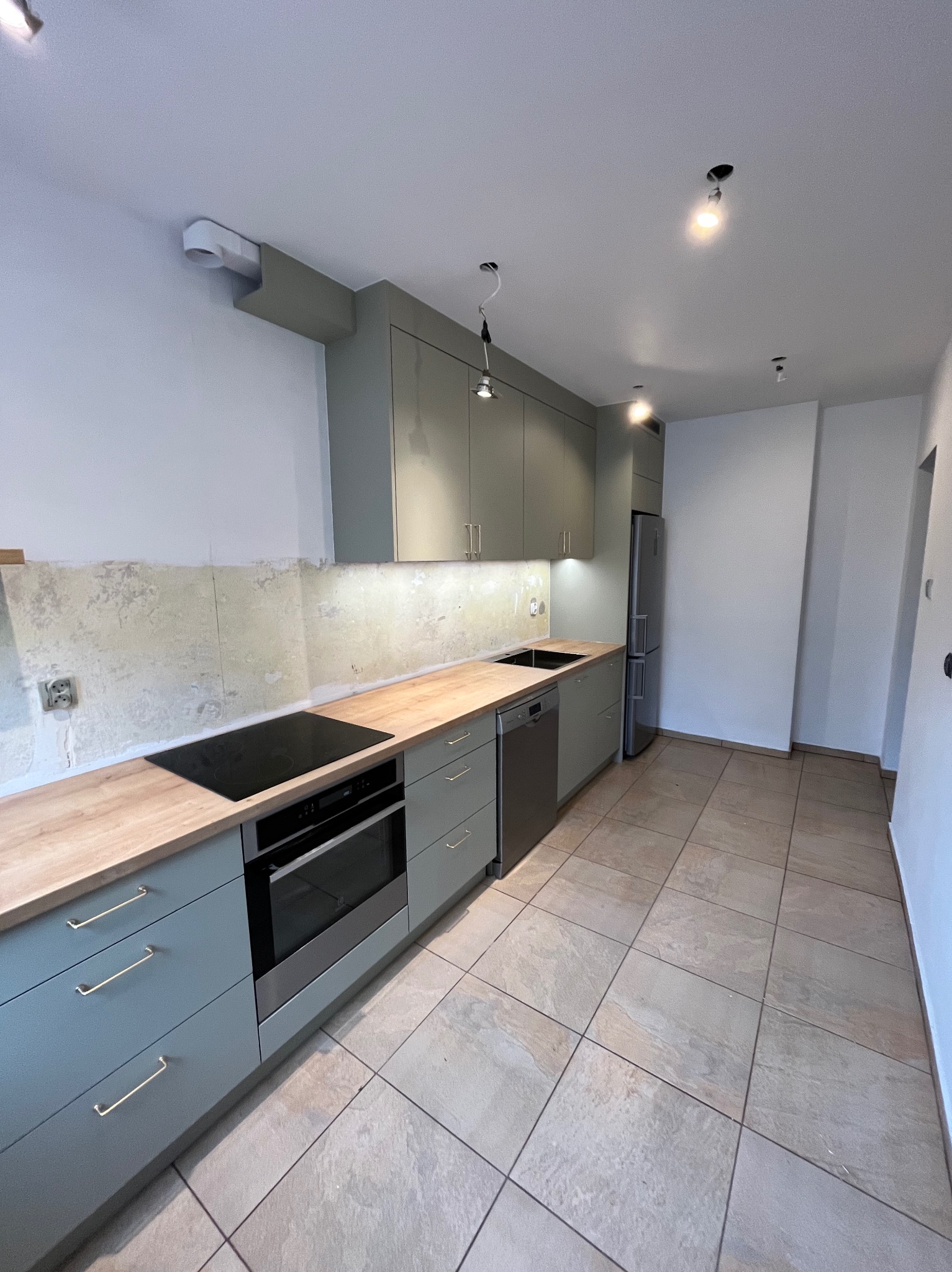 Nowoczesna kuchnia z szarymi szafkami i drewnianym blatem. Widoczne sprzęty AGD: piekarnik, płyta indukcyjna, zmywarka i lodówka. Ściana nad blatem przygotowana do wykończenia.