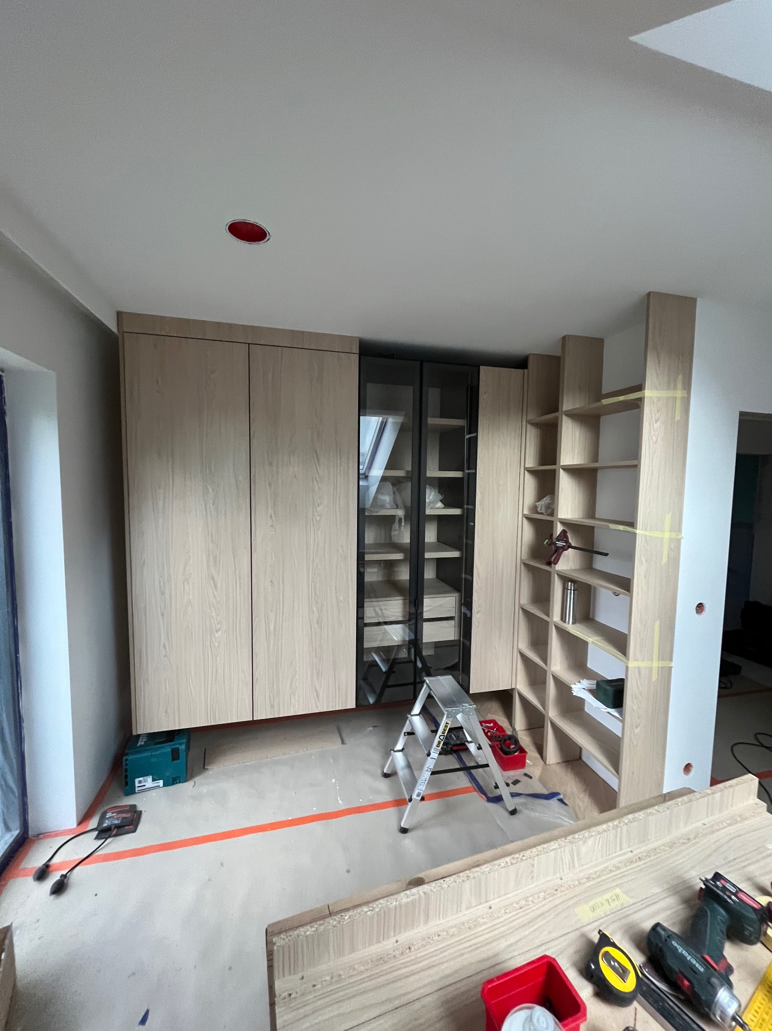 Wnętrze w trakcie prac: zabudowa meblowa z jasnego drewna, częściowo zamontowana szafa z przeszklonymi drzwiami, regał z półkami. Widoczne narzędzia i drabina.