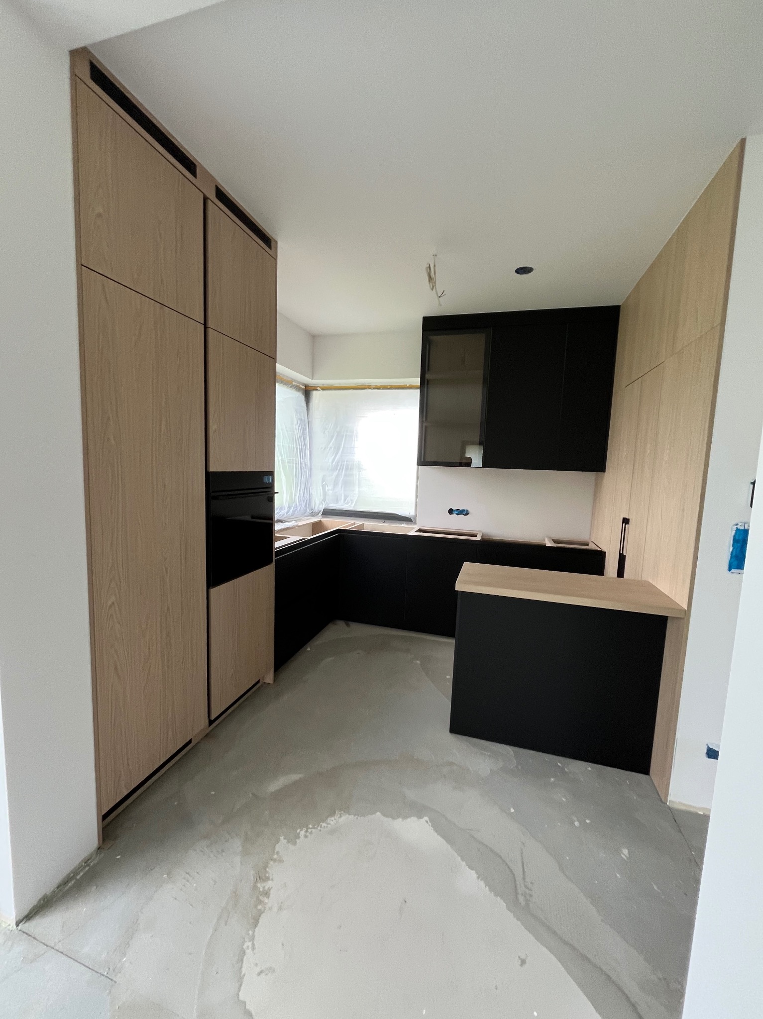 Częściowo zamontowana, nowoczesna kuchnia z czarnymi szafkami i drewnianymi panelami. Widoczne niedokończone podłogi i okno zabezpieczone folią.