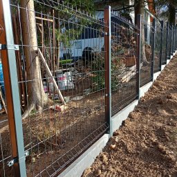 MASTER-BUD - Nowoczesne ogrodzenie panelowe z podmurówką betonową, widok z boku, z elementami krajobrazu w tle i wiaderkami z farbą przy słupku.