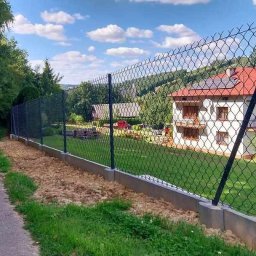 MASTER-BUD - Nowoczesne ogrodzenie z siatki, czarne słupki, betonowa podmurówka, widok na dom z czerwonym dachem i panele słoneczne w tle, zielona trawa i drzewa.