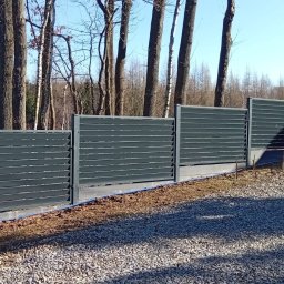 MASTER-BUD - Nowoczesne, poziome ogrodzenie panelowe w kolorze grafitowym, zamontowane na betonowej podmurówce. Widoczne drzewa w tle i żwirowe podłoże.