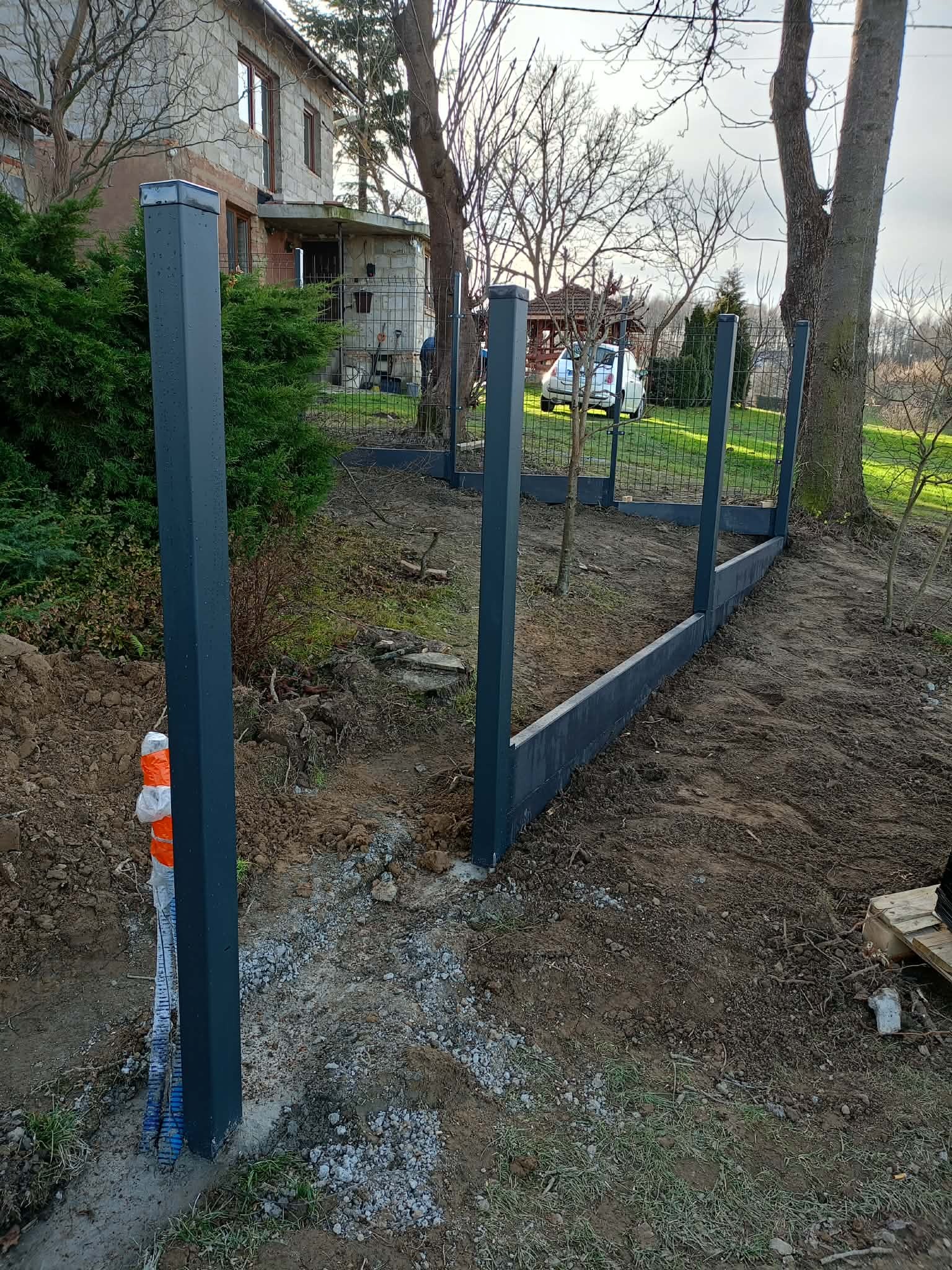 Wkopane słupki i podmurówka pod ogrodzenie panelowe na działce, widoczne narzędzia i częściowo zamontowana siatka w tle. Prace w toku, pochmurny dzień.